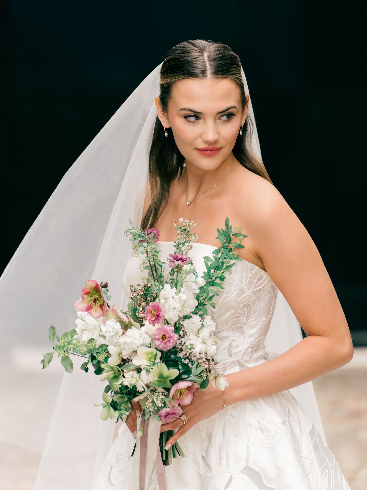 A bride in a white lace wedding dress poses with a romantic bouquet of pink and white flowers against a dark background.