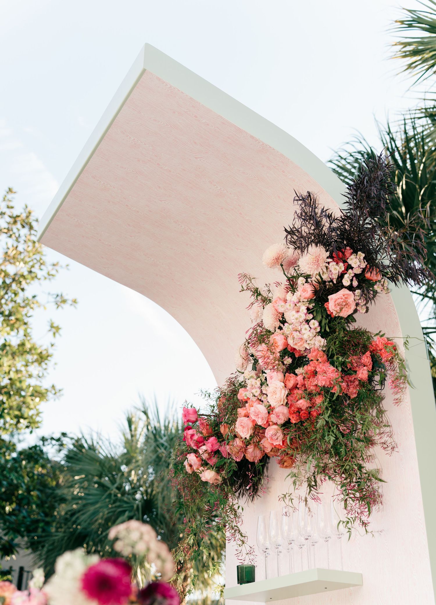Elegant white archway decorated with cascading coral and pink floral arrangements in a romantic outdoor garden setting.