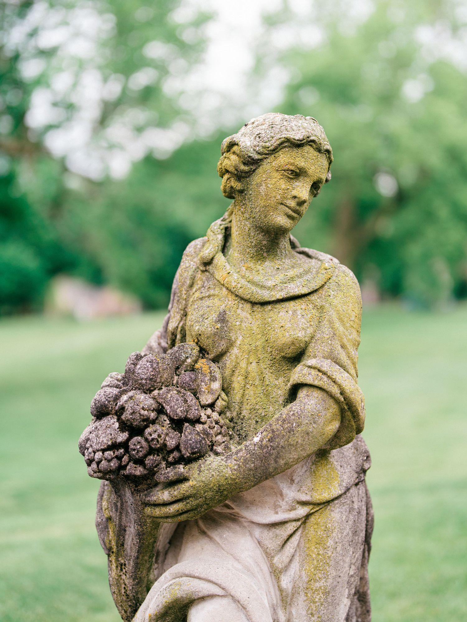 A weathered stone garden angel statue with moss patina holds flowers against a blurred green background.