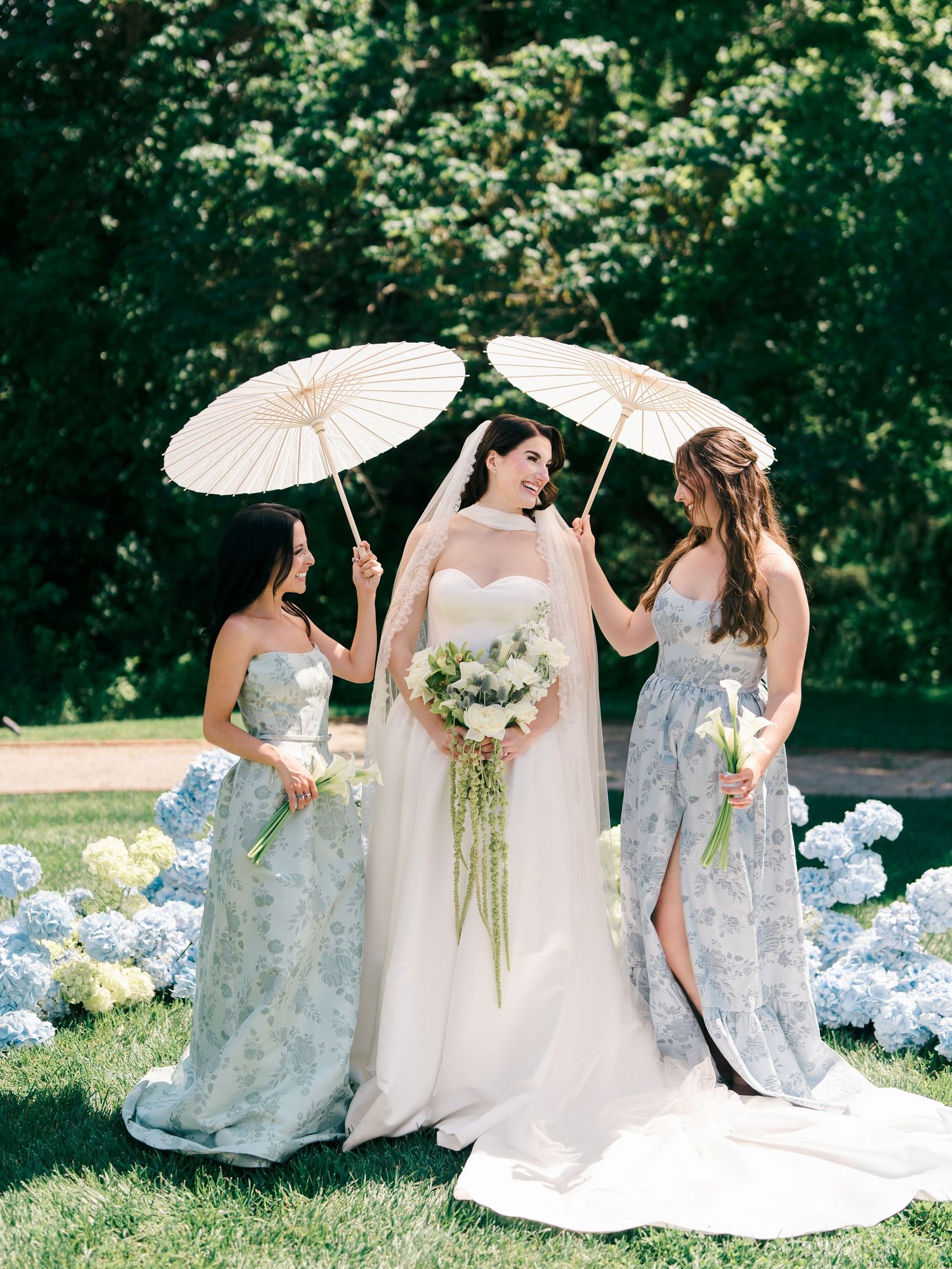 Bride in white gown with two bridesmaids in blue floral dresses holding parasols outdoors near hydrangeas.