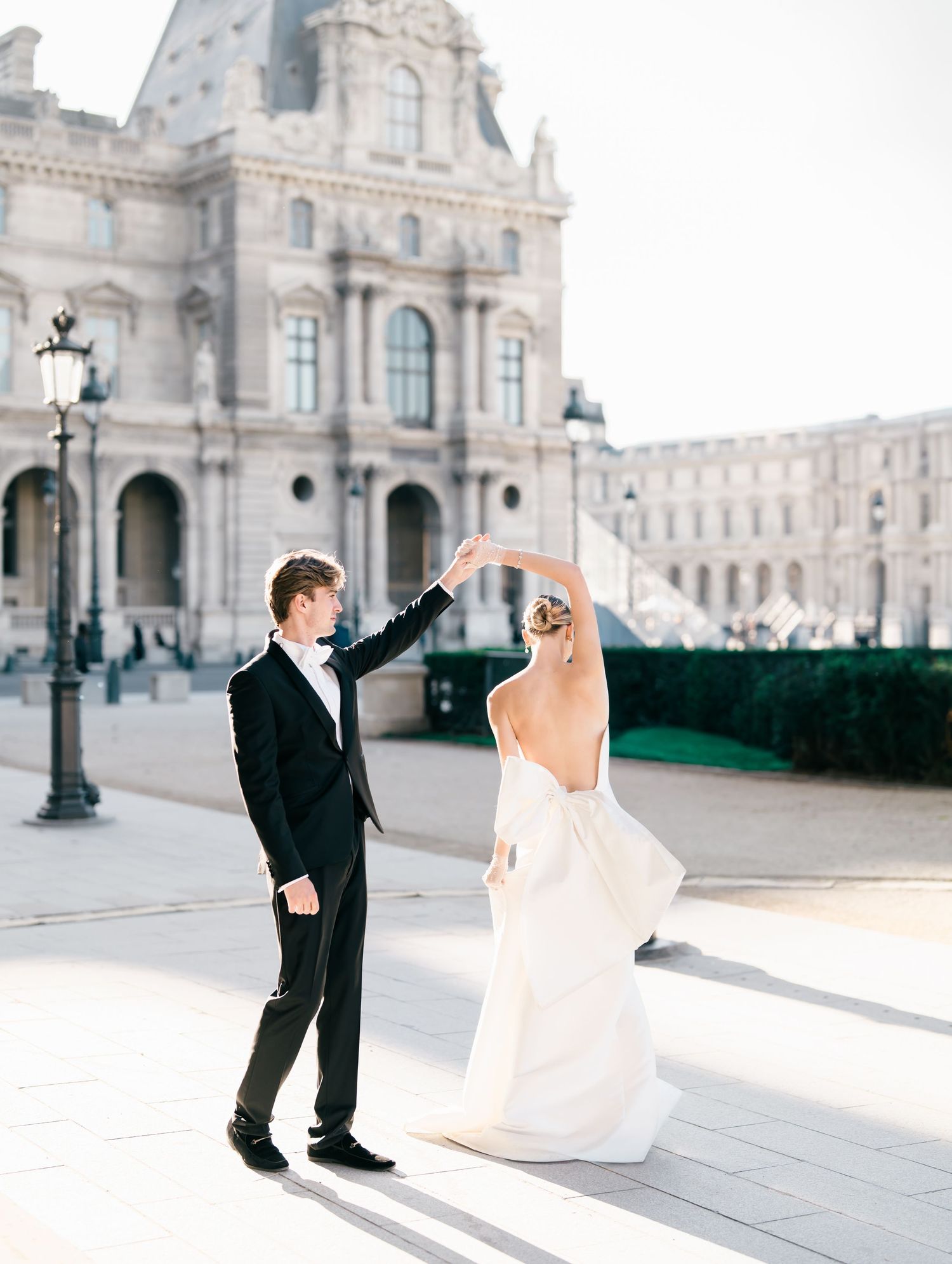 A romantic couple in formal attire dances elegantly in front of a grand European palace on a bright sunny day. The Louvre Museum in Paris.