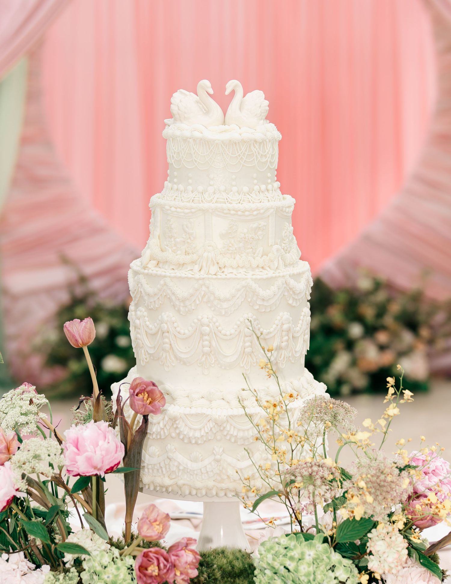 Elegant five-tier white wedding cake with lace details and swan topper, surrounded by pink and white floral arrangements.
