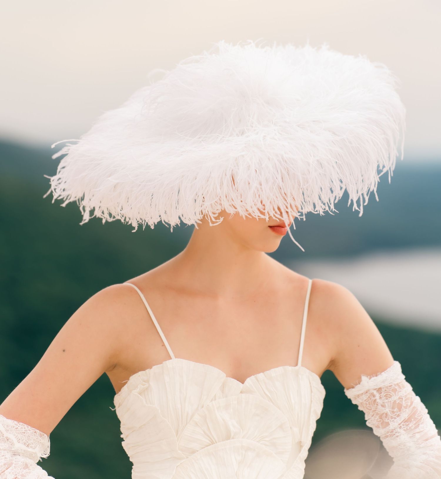 Bride wearing dramatic white feather wide-brim hat with strapless gown and lace gloves outdoors.