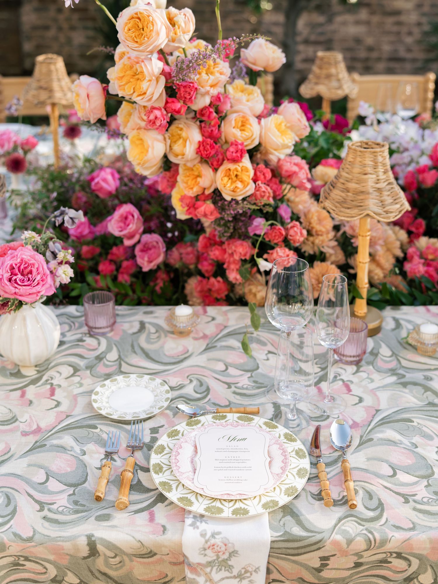 Elegant wedding table setting with pink and peach roses, crystal glassware, and a swirling patterned tablecloth.