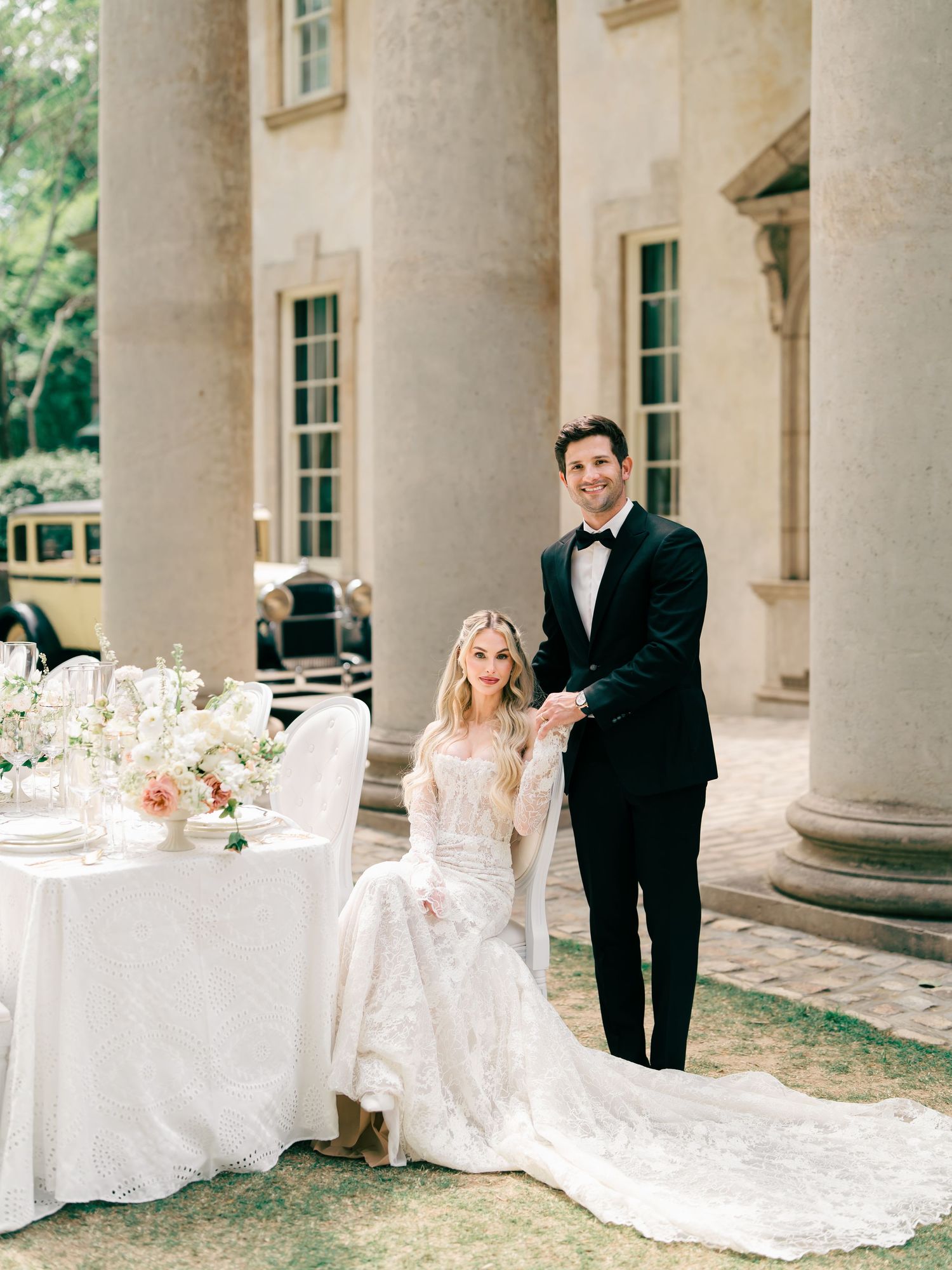 Elegant bride in lace gown seated beside groom in tuxedo at outdoor wedding reception near classical stone mansion.