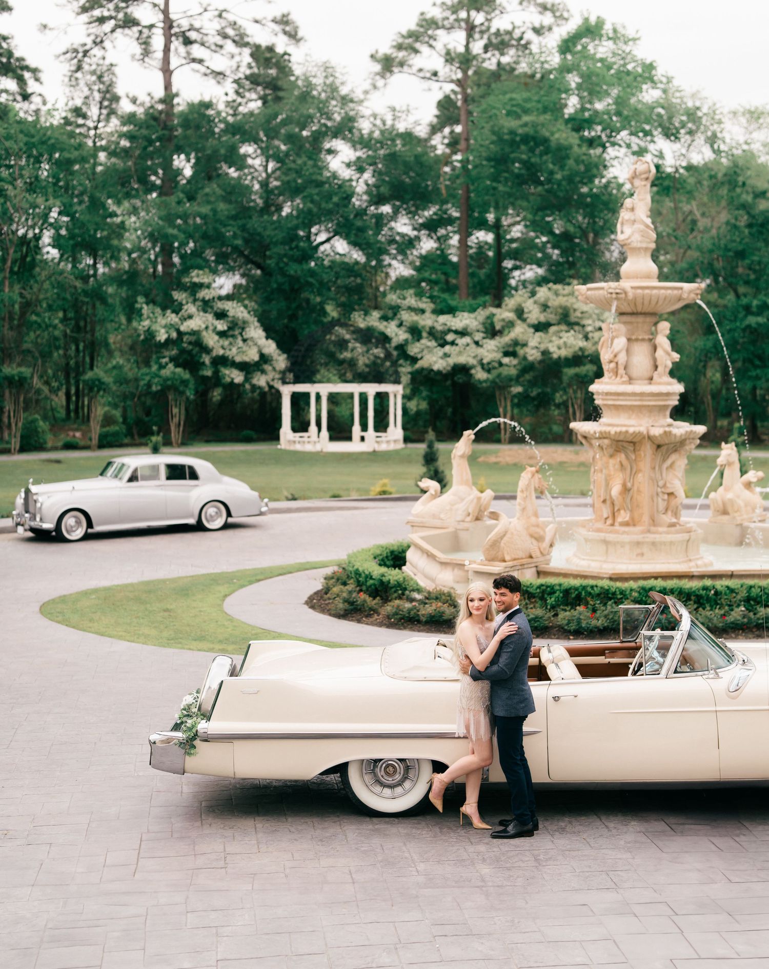 Vintage wedding photoshoot with classic cars and fountain in an elegant outdoor garden setting.