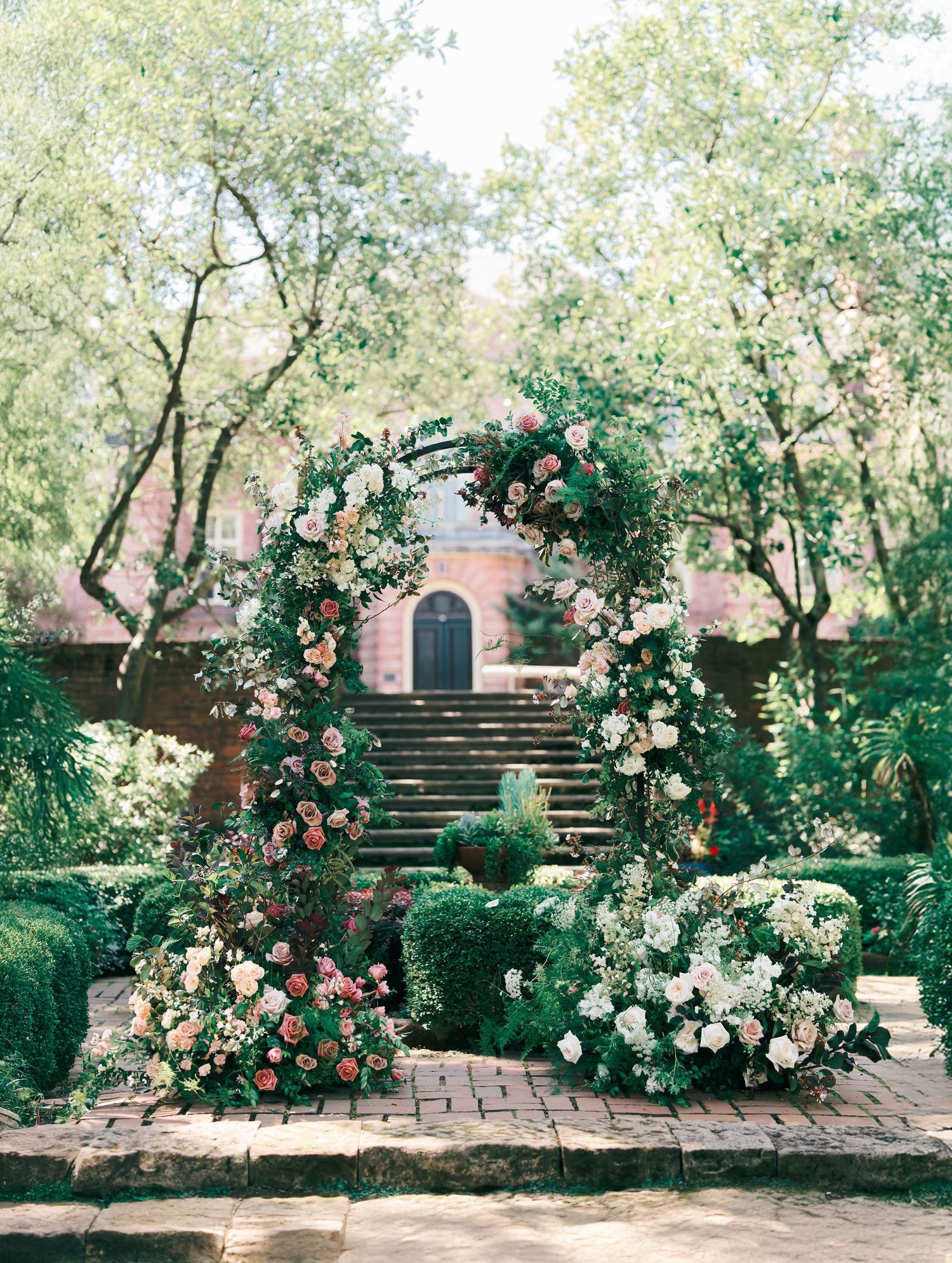 Beautiful garden path with pink climbing roses leading to a brick entryway with curved stone steps and lush greenery.