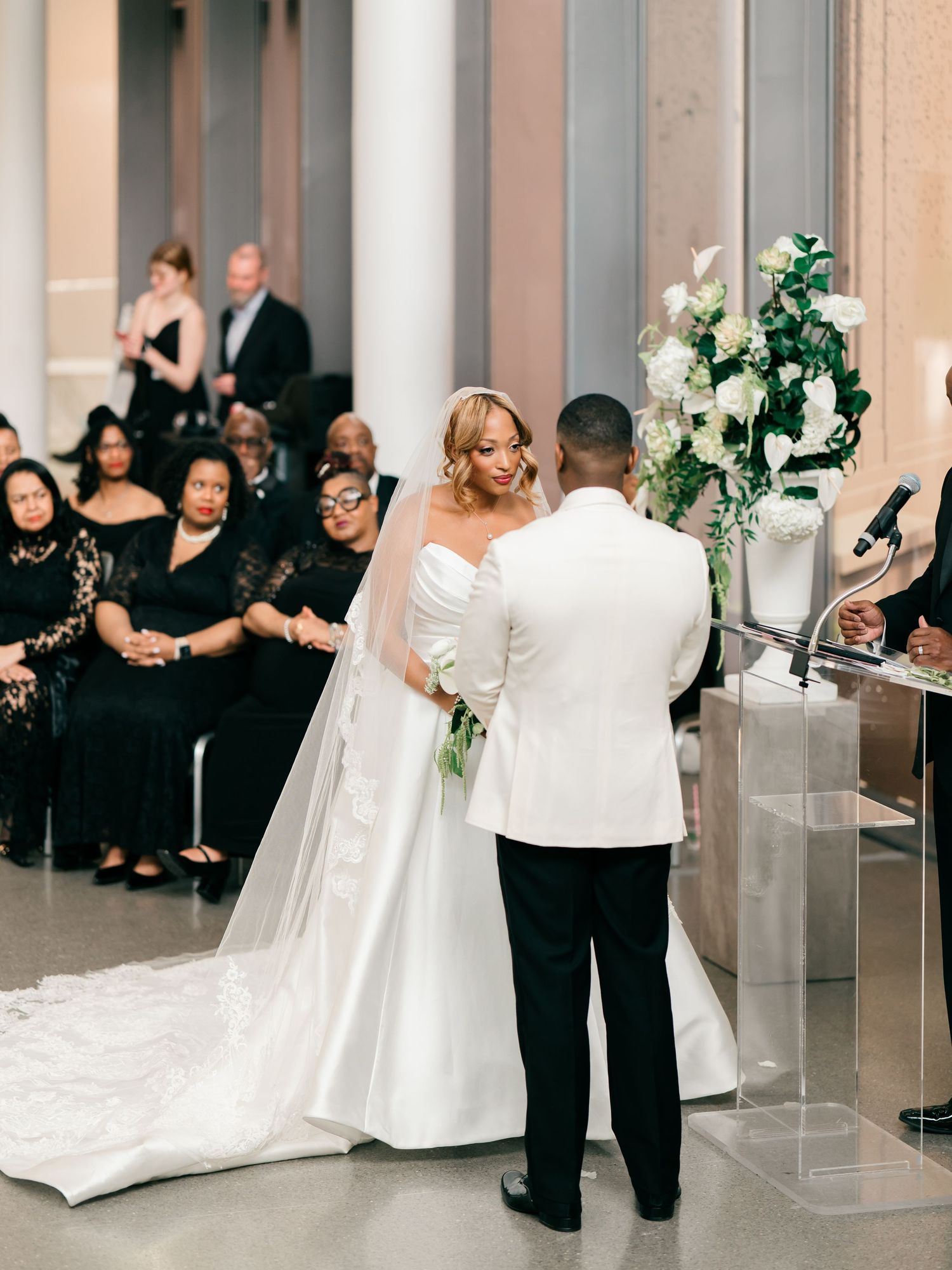 Wedding party dances in a grand white ballroom with elegant floral decor and classic columns.