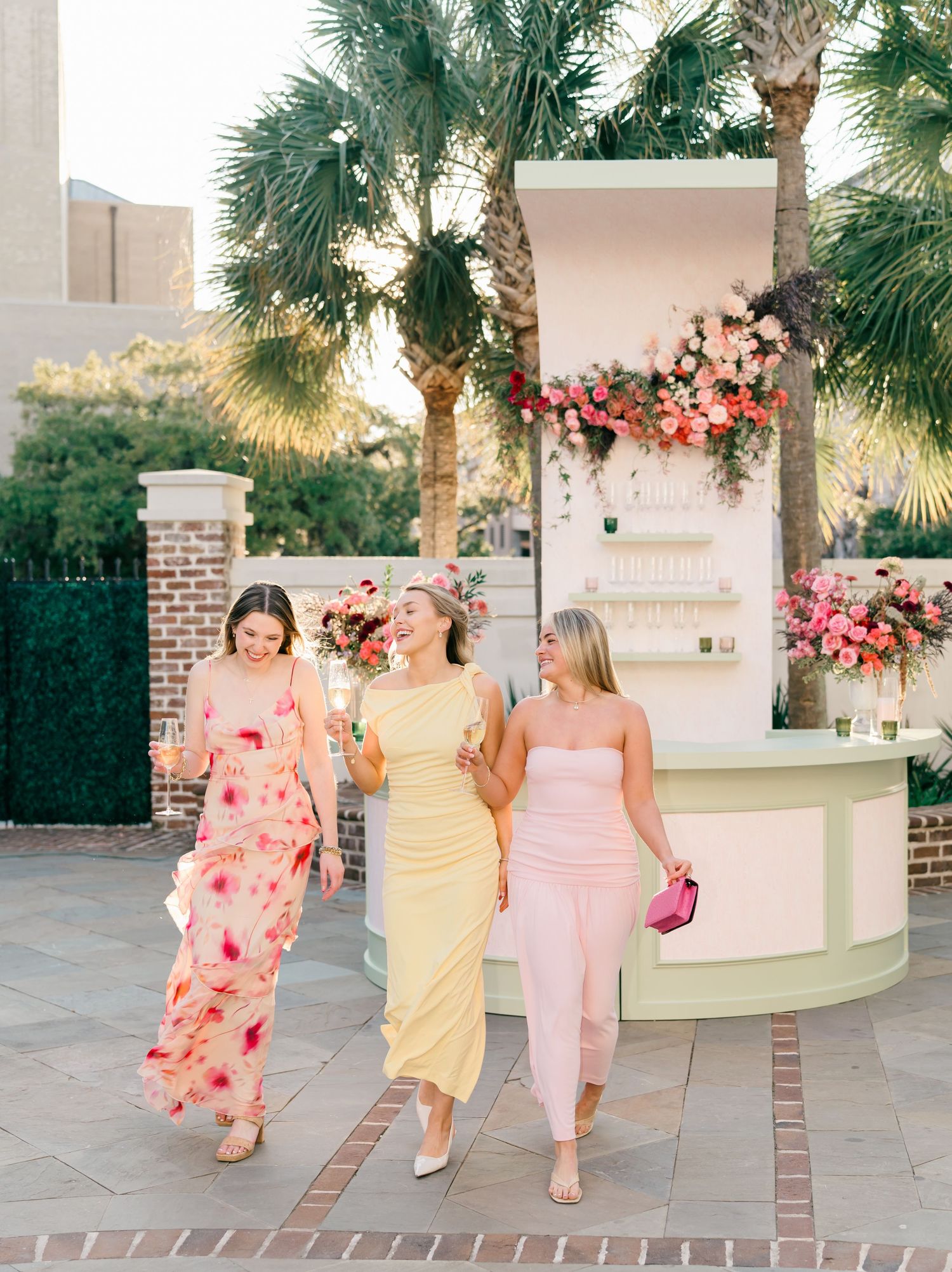 A group in coordinating pink and yellow dresses walking together on a brick pathway near palm trees at sunset.