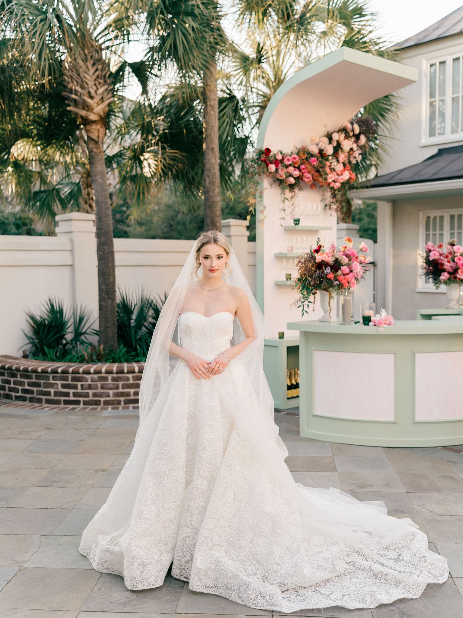 Elegant white lace wedding dress displayed in a southern garden setting with palm trees and pink floral decor.