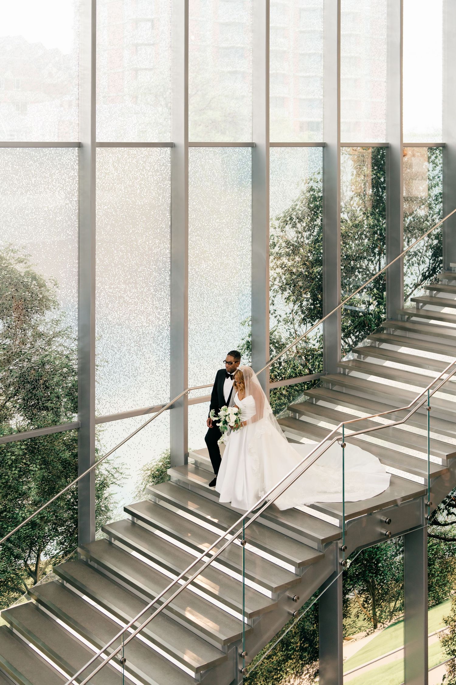 A couple in formal wedding attire descends a modern staircase with natural light streaming through large windows.
