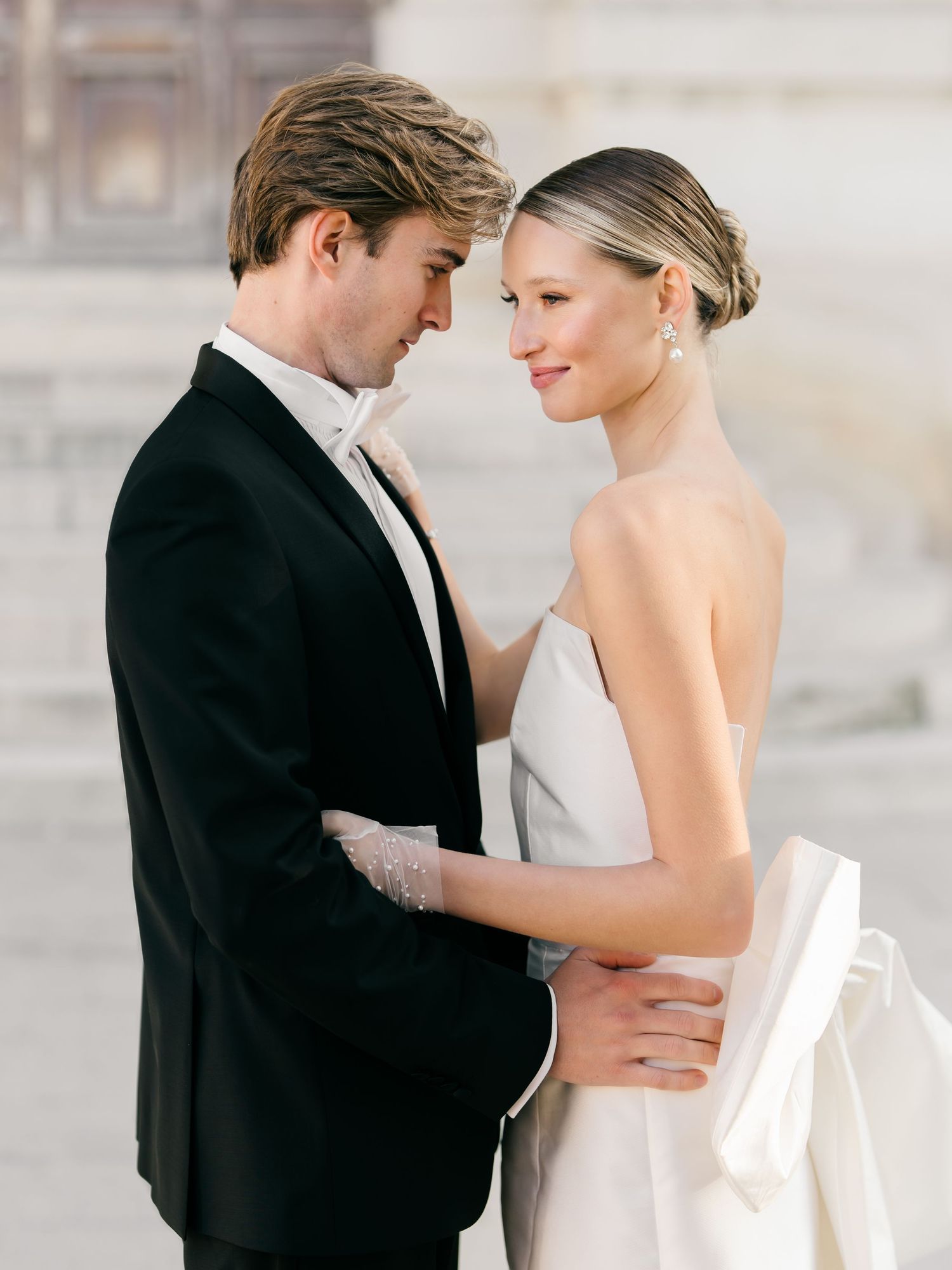 A romantic wedding couple embraces in formal attire while sharing an intimate moment in a soft, light-filled setting. The Louvre, Paris.