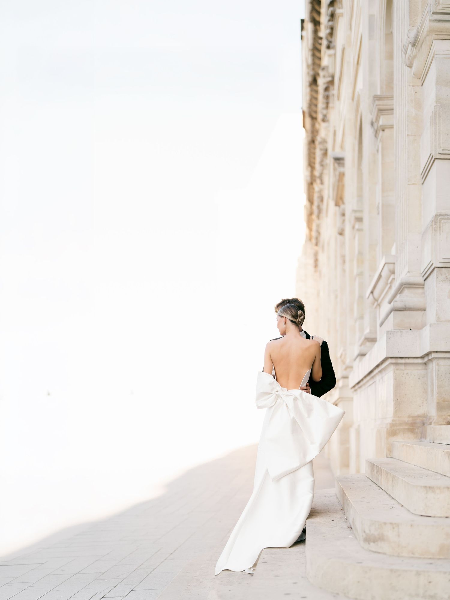 Bride in backless white gown with bow stands beside ornate stone building, elegant destination wedding photography. At the Louvre in Paris.