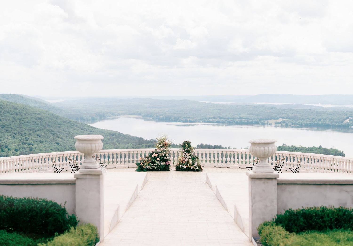 Elegant stone terrace with white balustrades overlooking a serene river and lush green mountains under a cloudy sky.