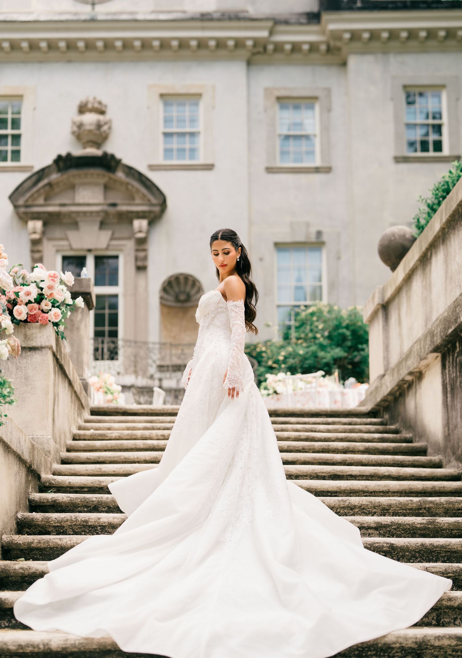 Bride in elegant white gown with long train poses on stone steps of a grand manor house.