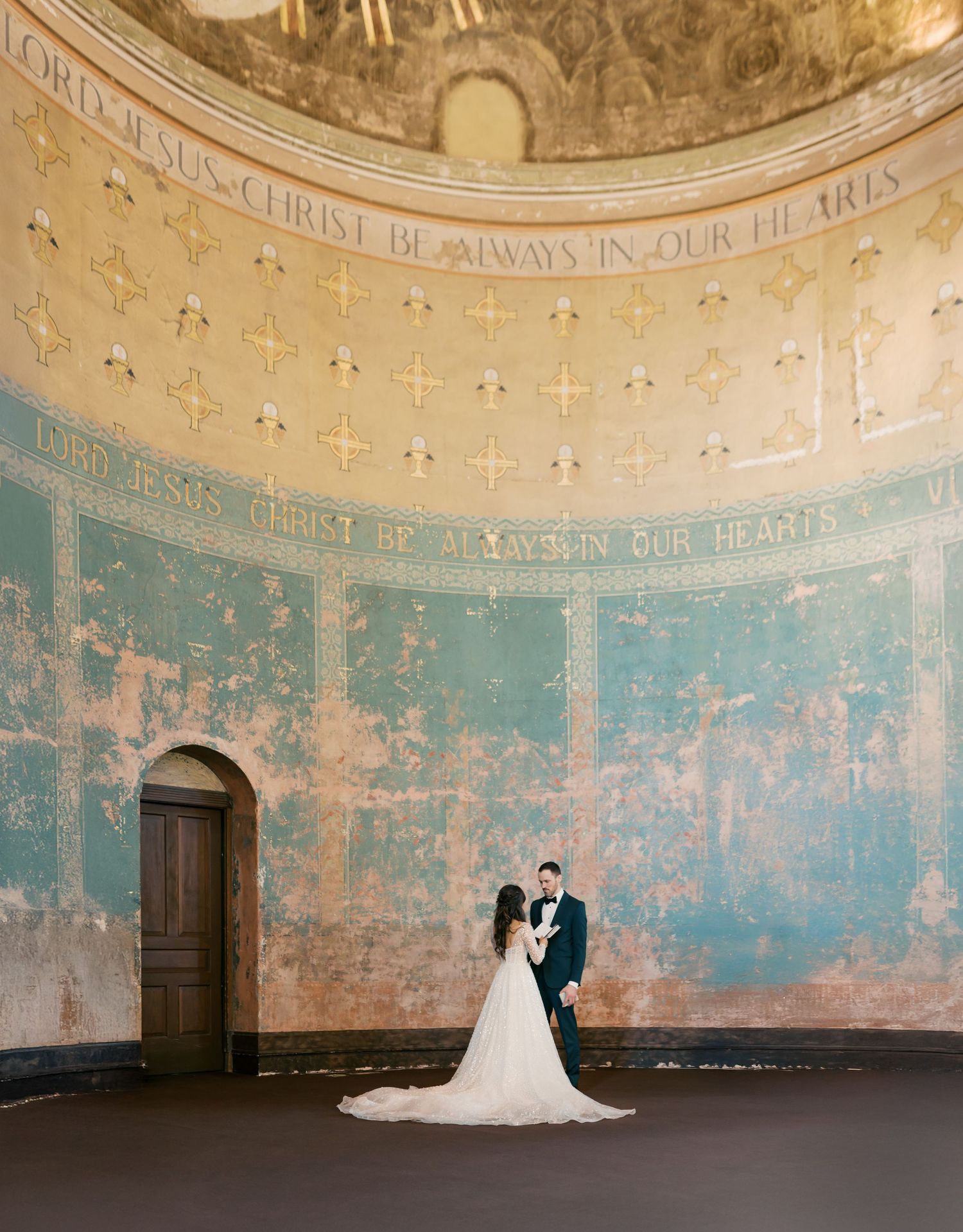 A couple stands in a circular room with distressed turquoise walls, ornate ceiling details and a classic wooden doorway.