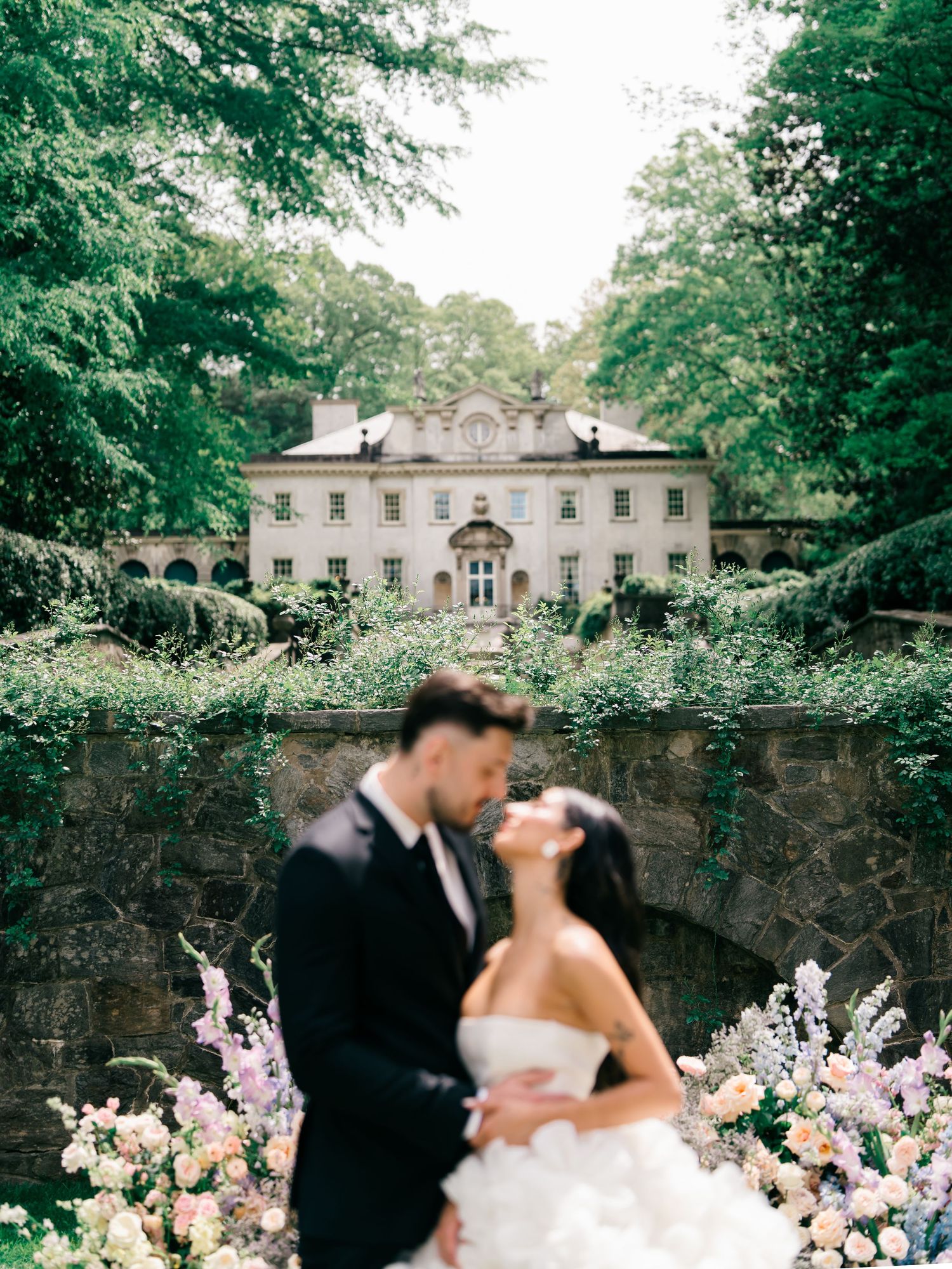 Couple shares romantic moment in front of elegant ivy-covered mansion surrounded by lush gardens and pastel floral arrangements.