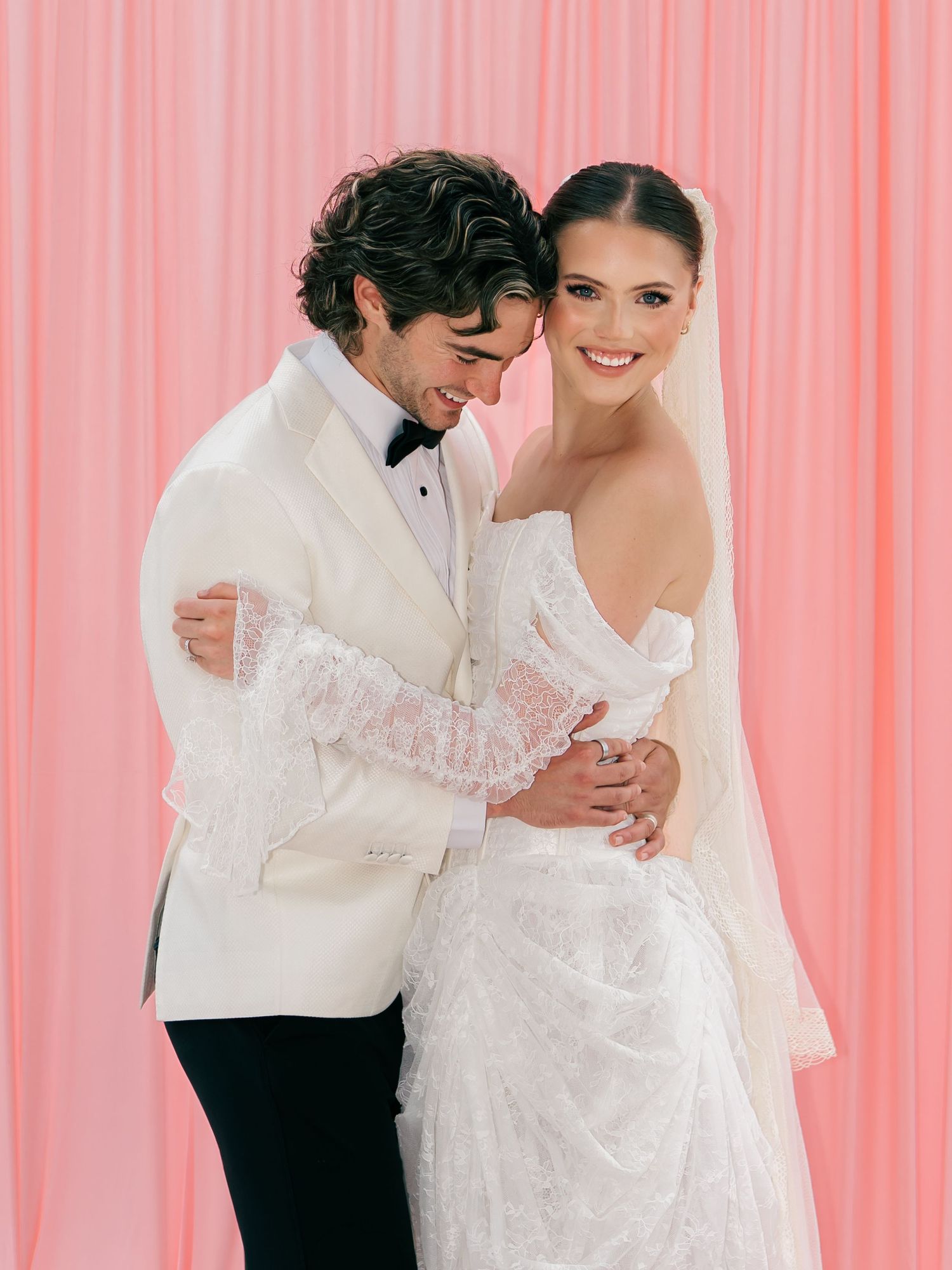 Bride and groom in elegant wedding attire pose together against a soft pink backdrop, smiling joyfully.