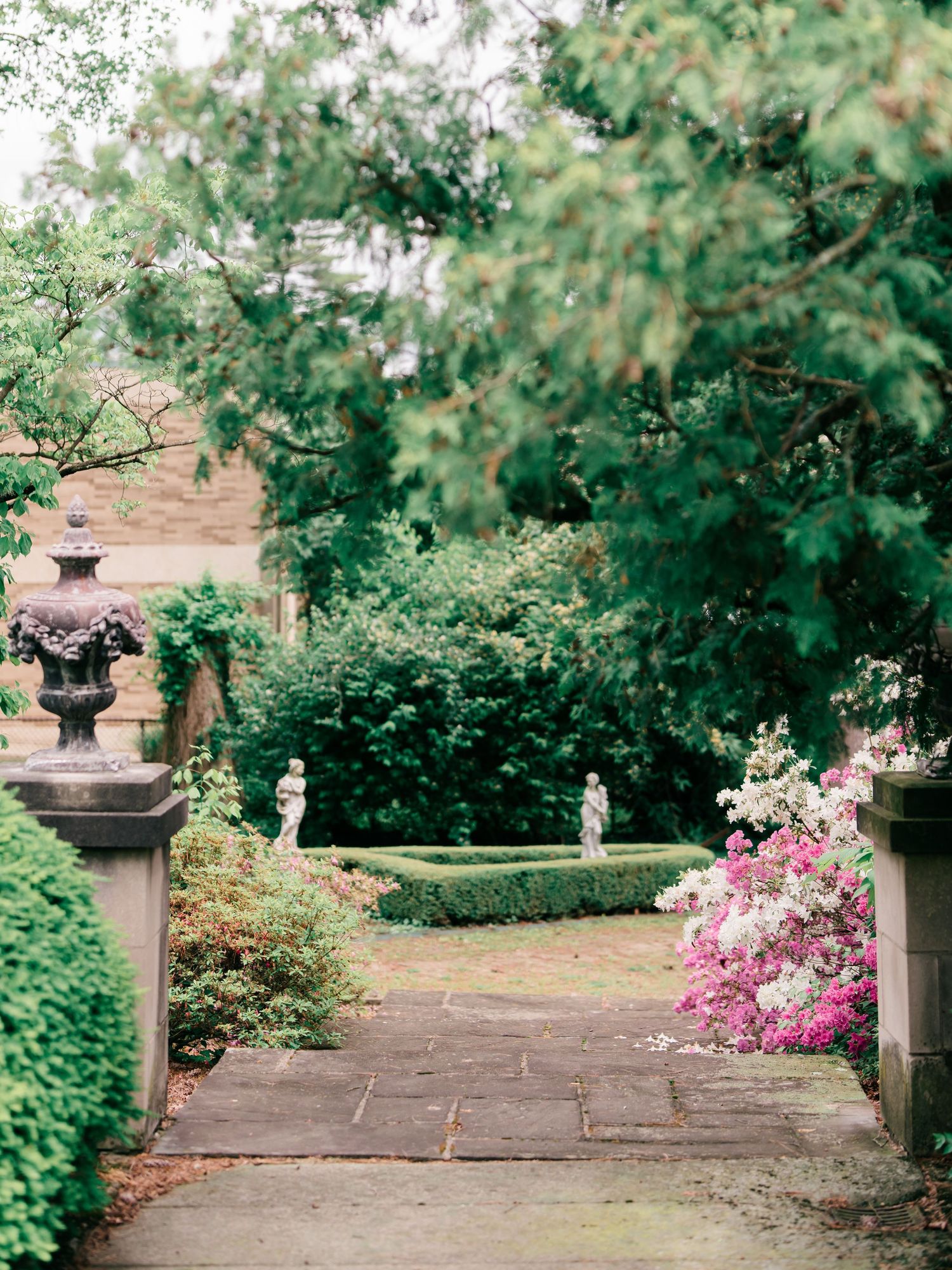 A stone-paved garden path lined with ornate pillars, statues, and pink azaleas leads through a lush green landscape.
