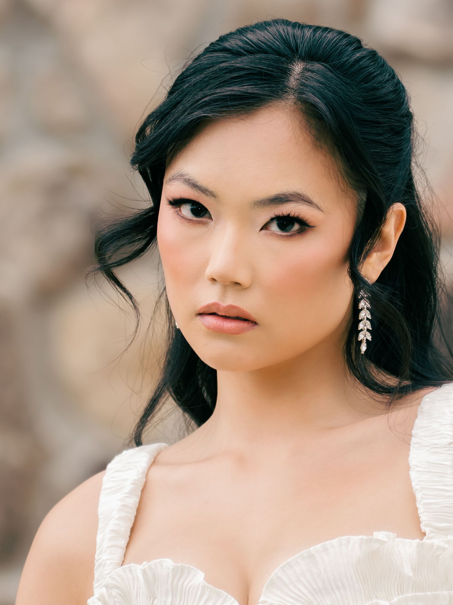 Elegant bride with dark updo hair, dramatic eye makeup, and silver leaf drop earrings wearing a white strapless gown.