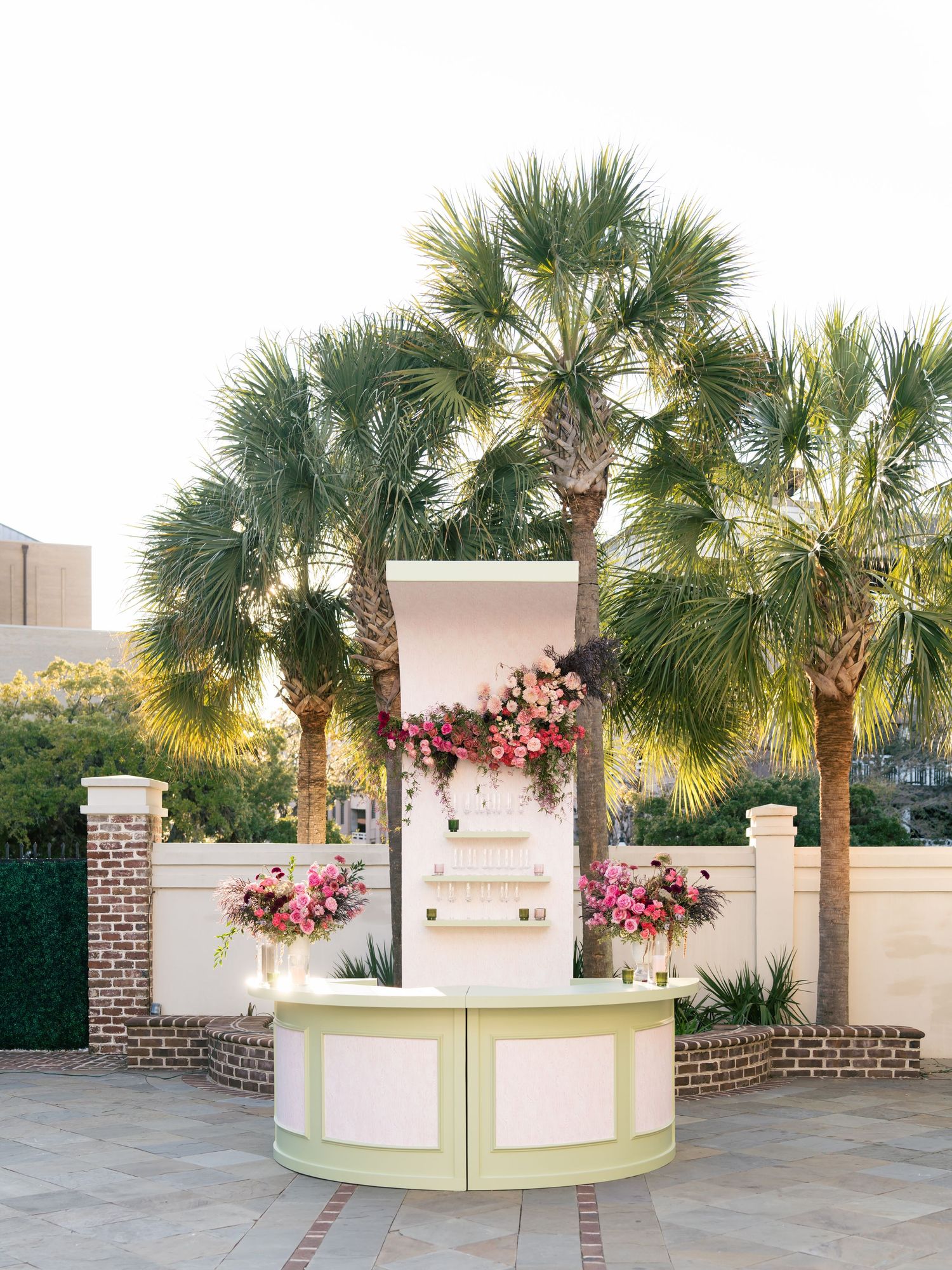 Elegant outdoor wedding ceremony setup with palm trees and pink floral arrangements on white pedestals at sunset.
