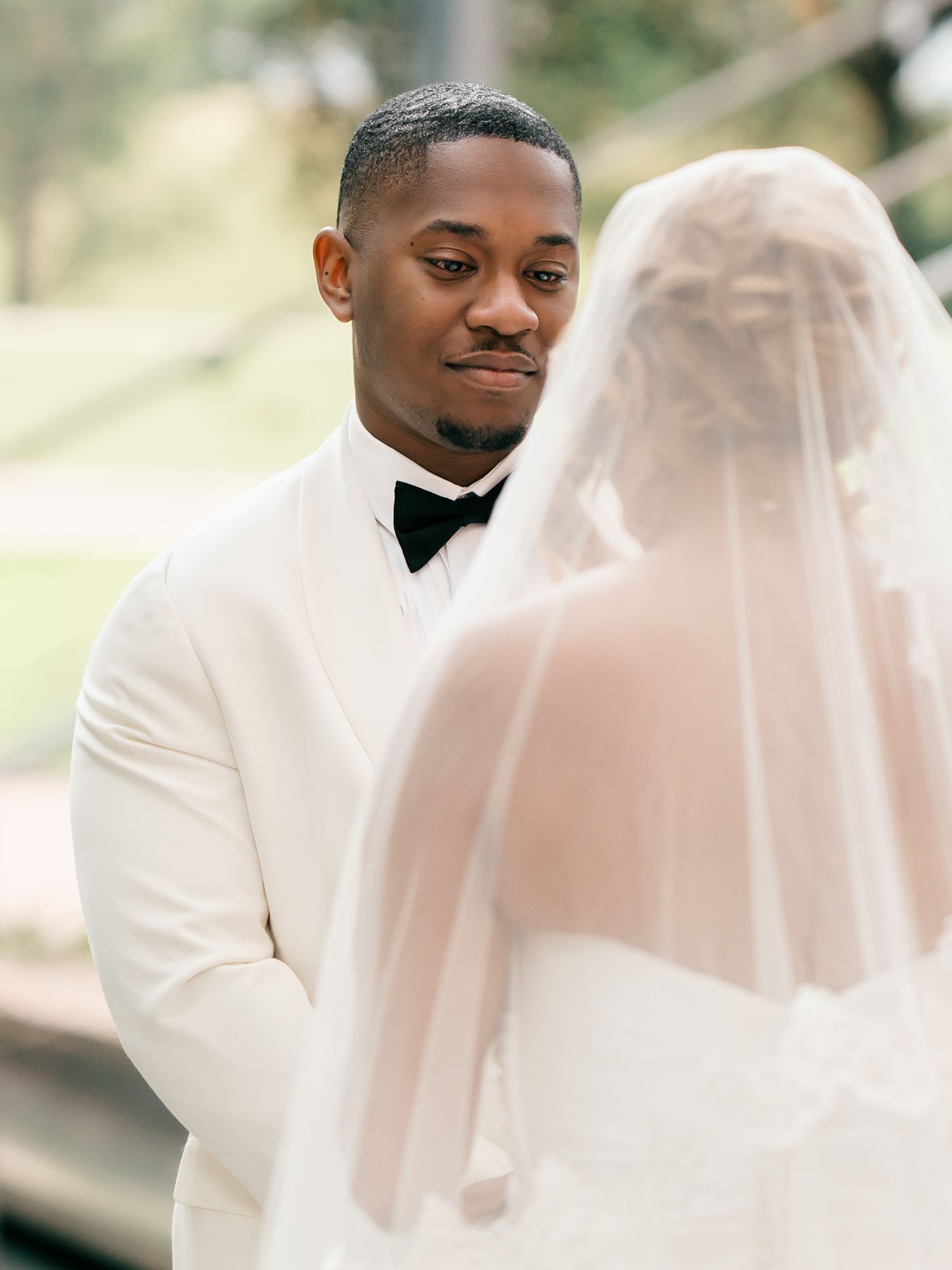 A groom in a white tuxedo jacket and black bowtie gazes lovingly at his bride in her flowing veil during their wedding ceremony.