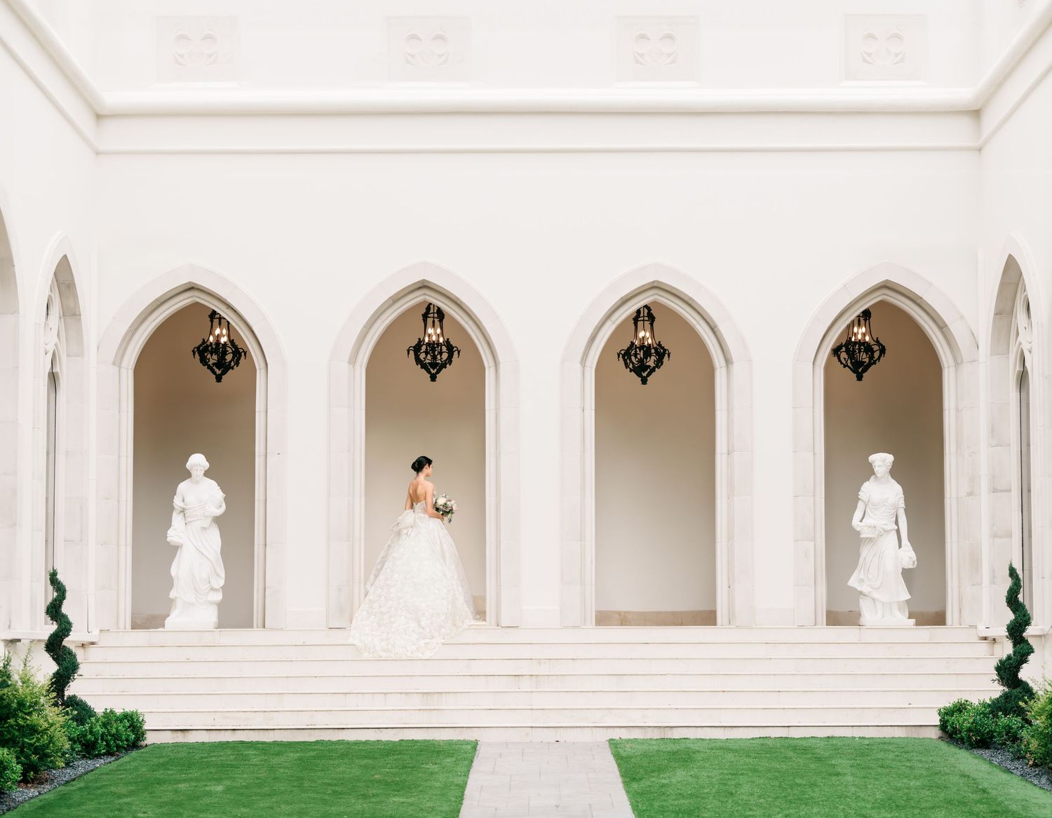 White classical colonnade with arched niches and decorative statues creates an elegant architectural walkway in a garden setting.