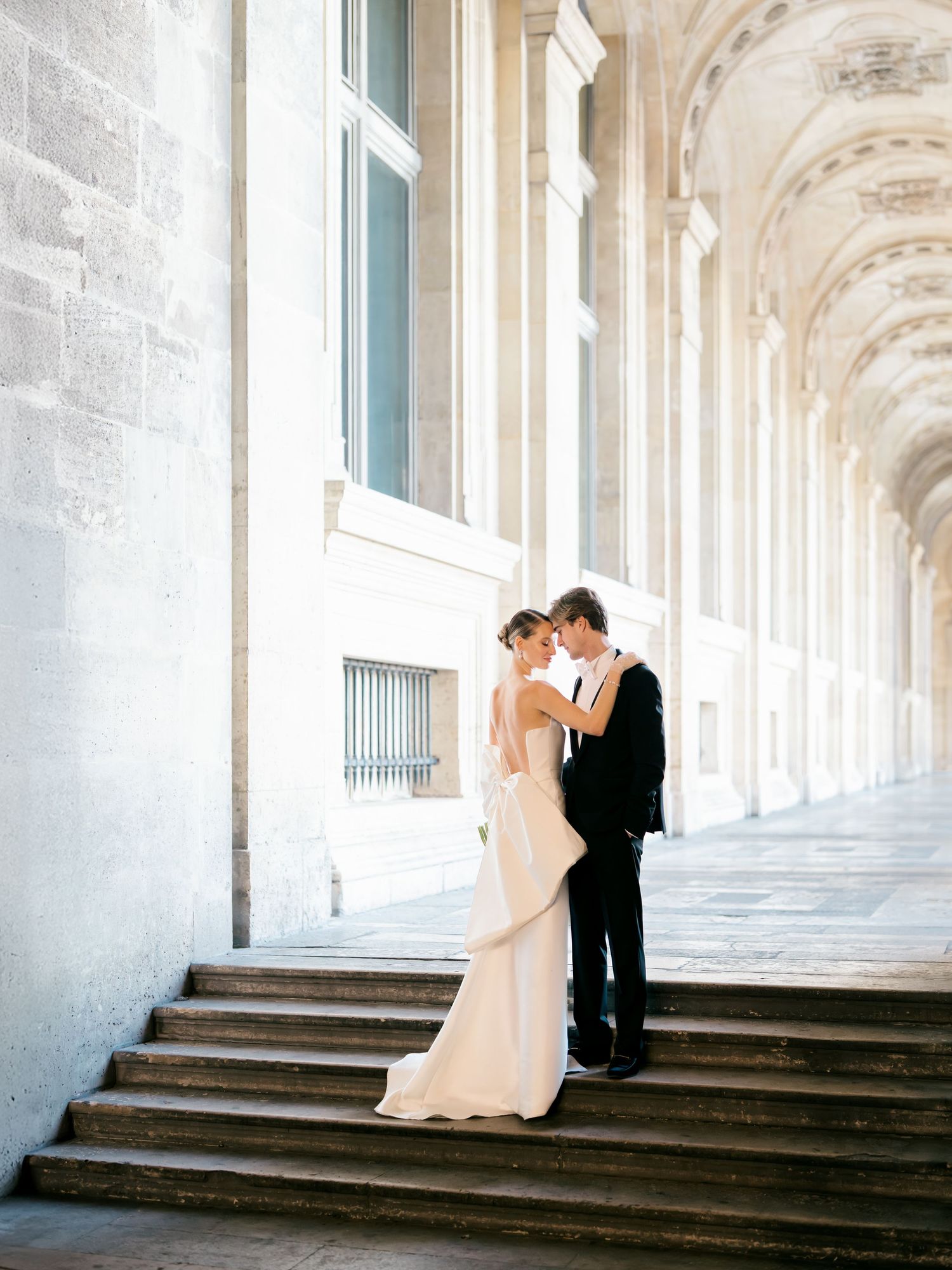 A romantic couple in formal wedding attire share an intimate moment in a grand columned hallway at The Louvre Museum in Paris, France.