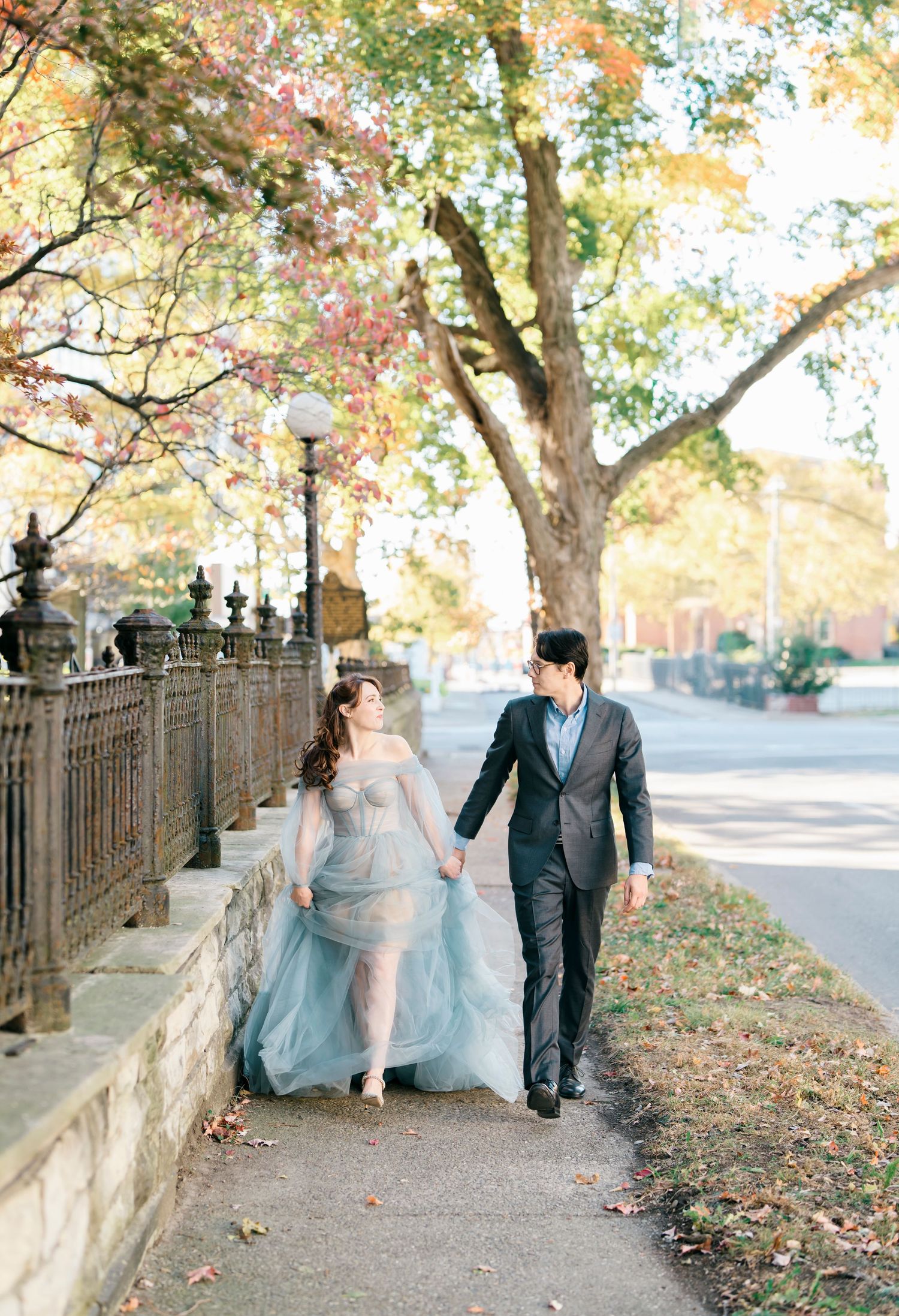 A couple in wedding attire walks along a tree-lined path during autumn with fallen leaves on the ground.