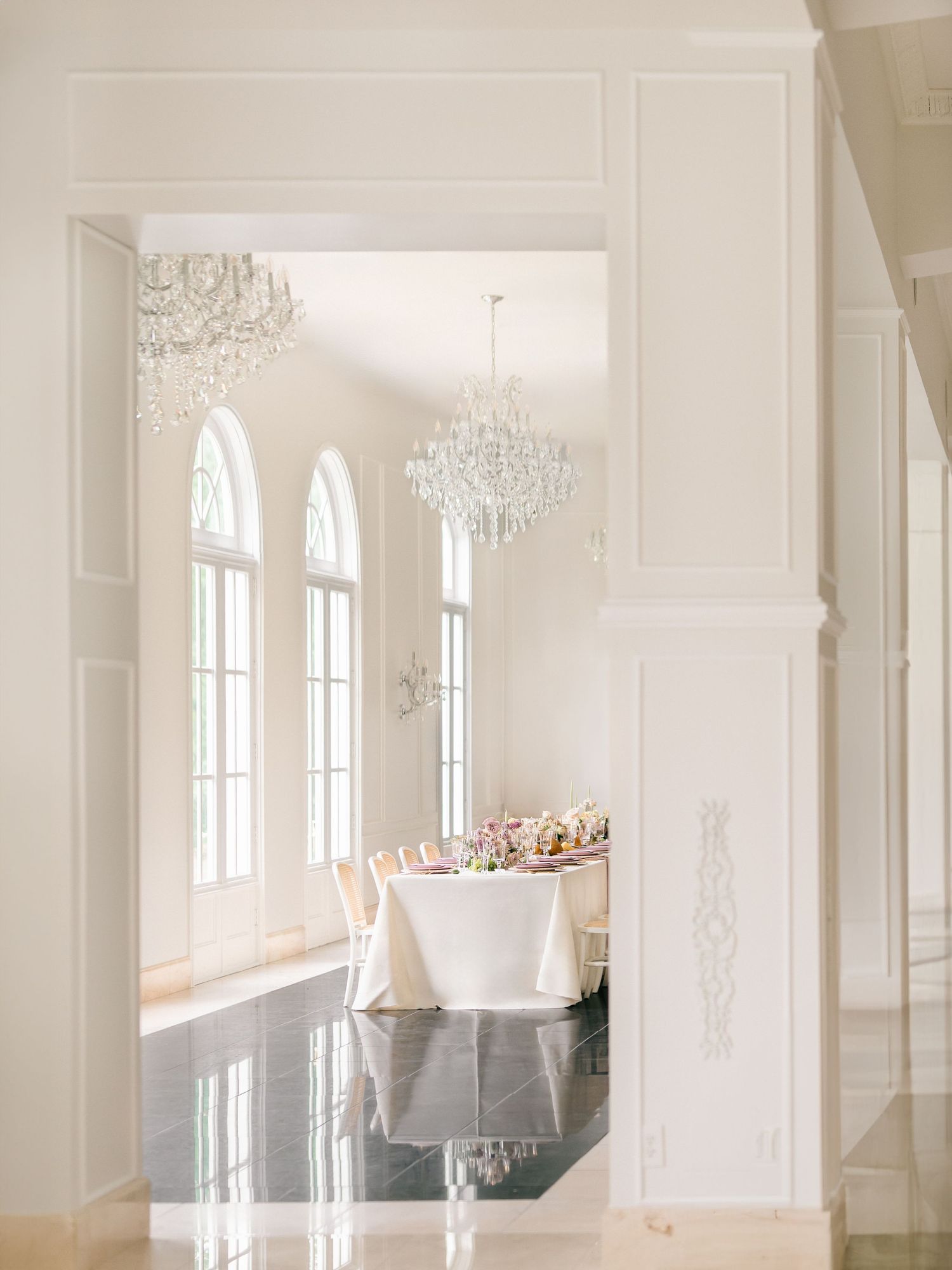 Elegant white ballroom with arched windows, crystal chandeliers and a decorated table setting in soft natural light.