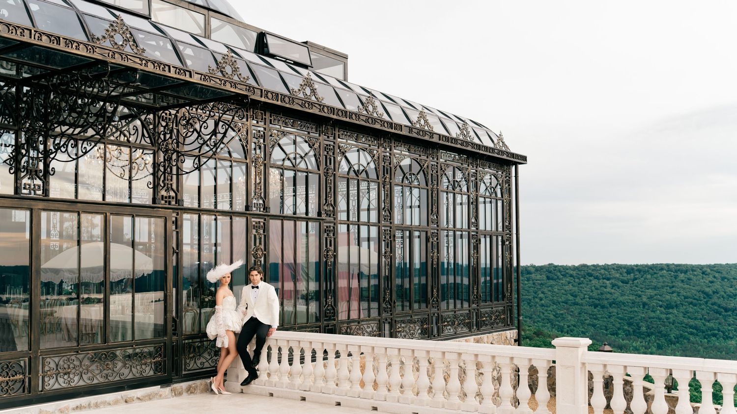 Couple embracing on a terrace beside an ornate Victorian glass greenhouse with lush green landscape views.