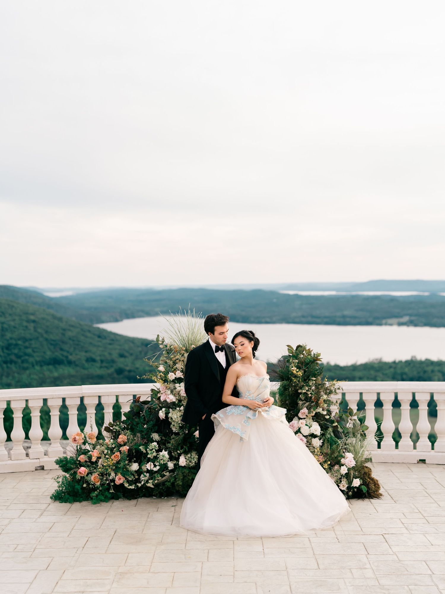 Bride and groom pose on elegant terrace with lush floral arrangements and scenic lake views in background.