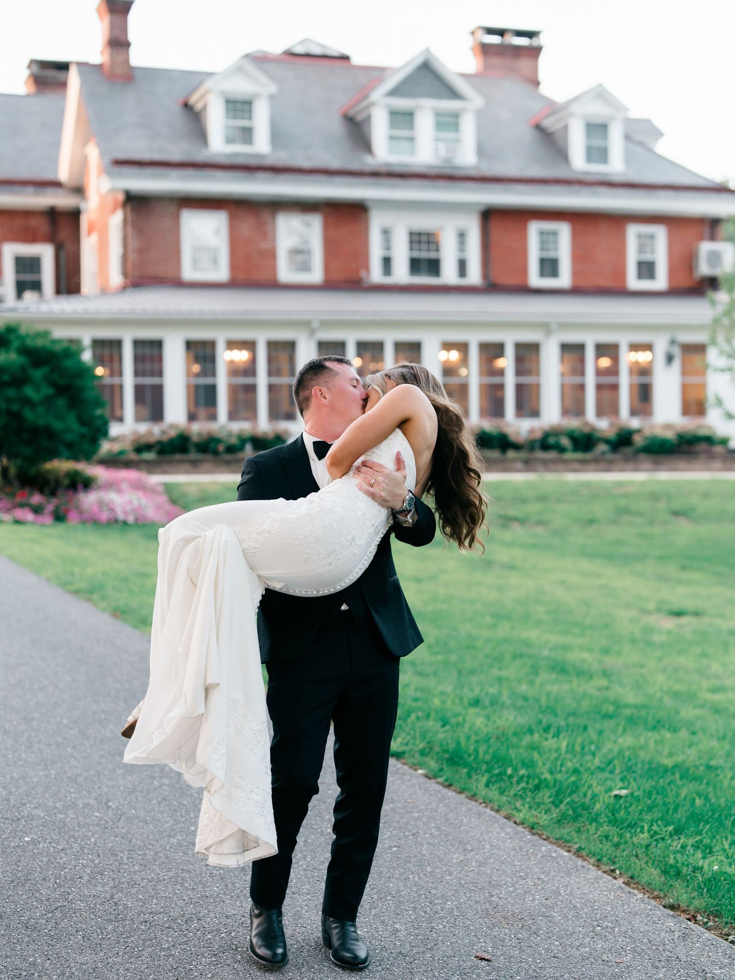 Groom lifts bride in white gown for a romantic kiss outside elegant brick mansion during golden hour wedding. The Cameron Estate.
