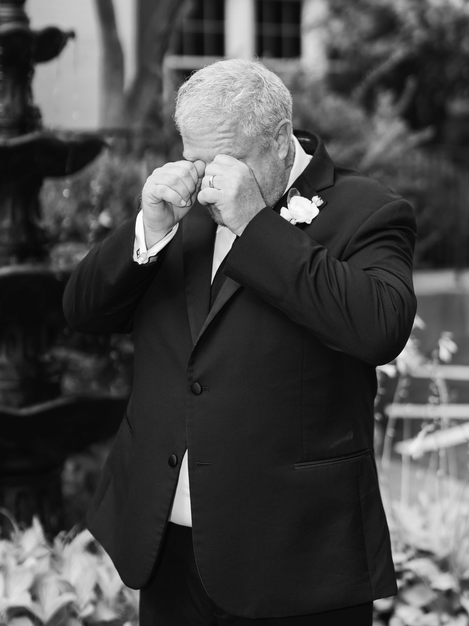 Emotional groom wiping tears at outdoor wedding ceremony, wearing black tuxedo with white boutonniere, fountain in background.