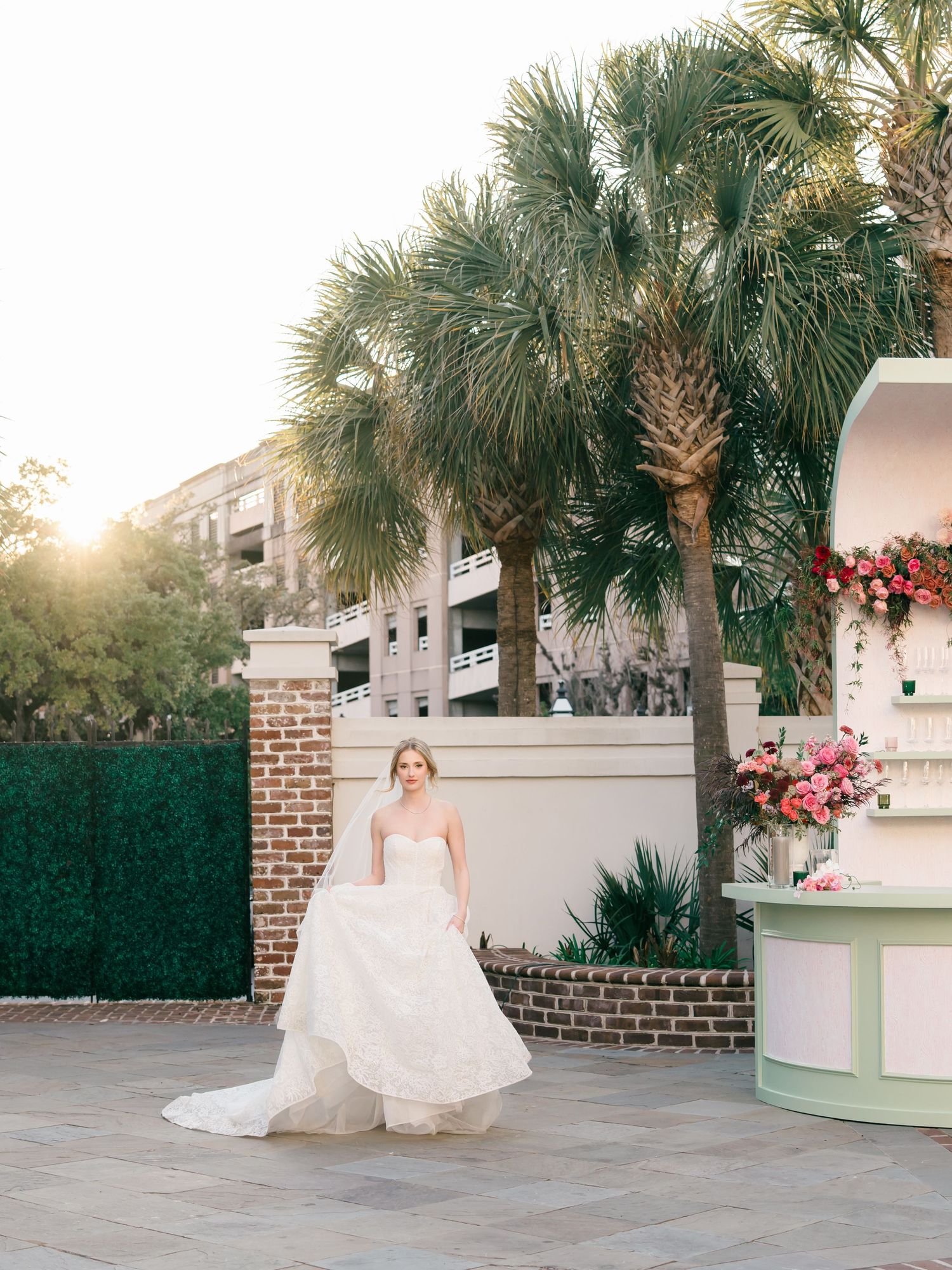 A bride in a flowing white gown twirls gracefully on a stone patio surrounded by palm trees at sunset.