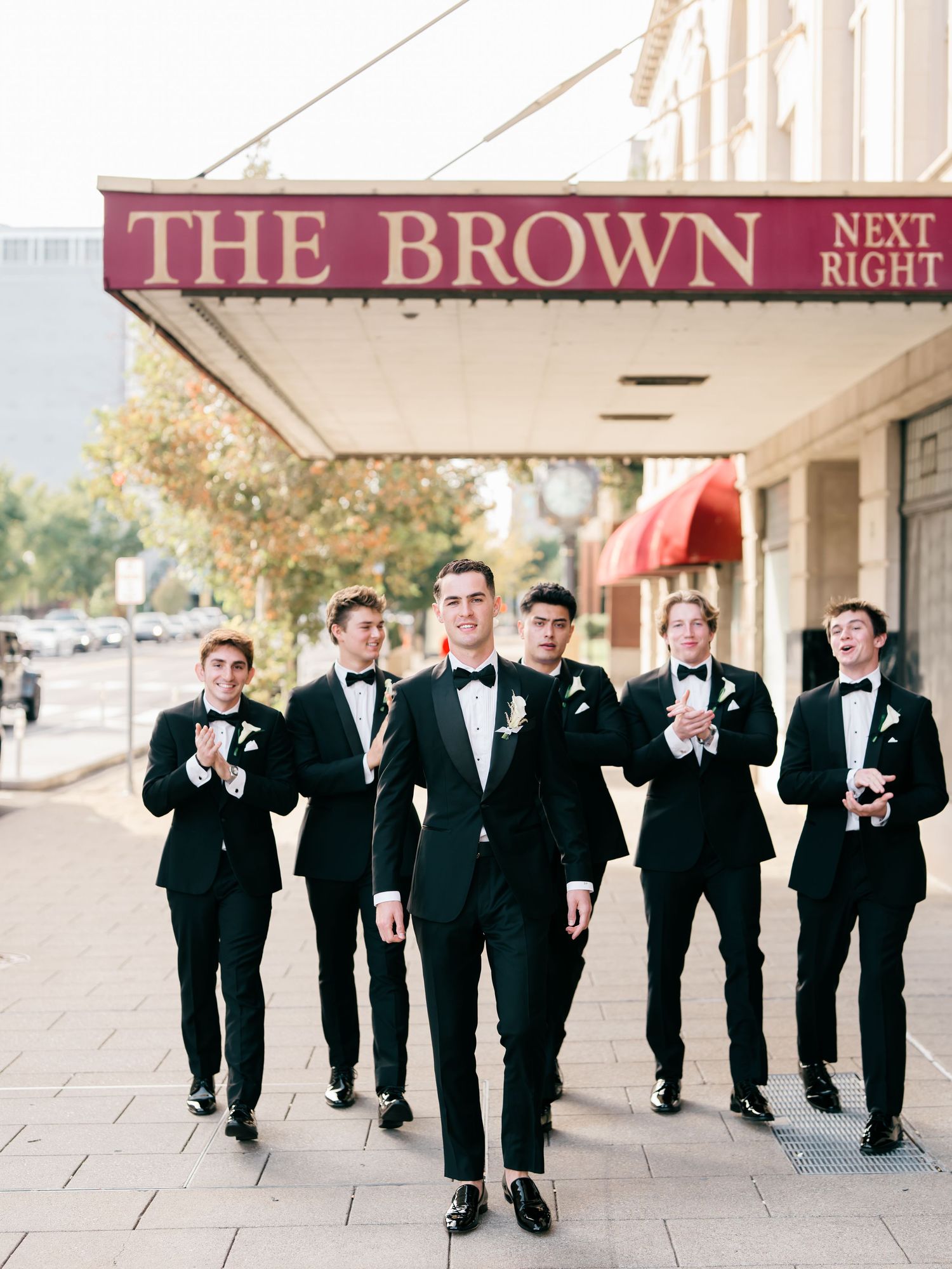 Groom & groomsmen, in black tuxedos and bow ties, walk together on a sidewalk beneath The Brown Hotel marquee. Louisville, Kentucky.