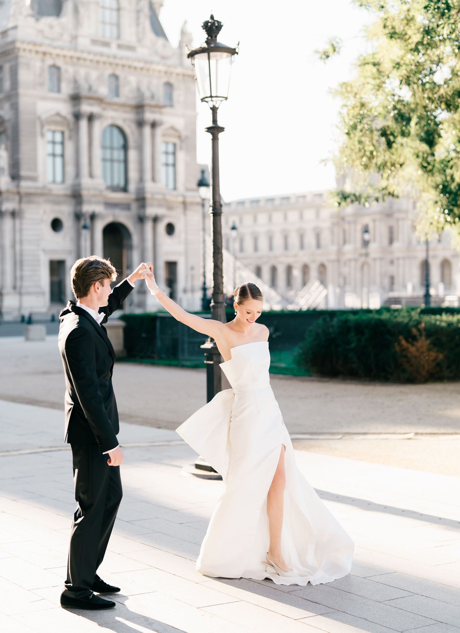 A couple in wedding attire dances gracefully in front of an elegant European palace at sunrise. The Louvre Museum Paris, France.
