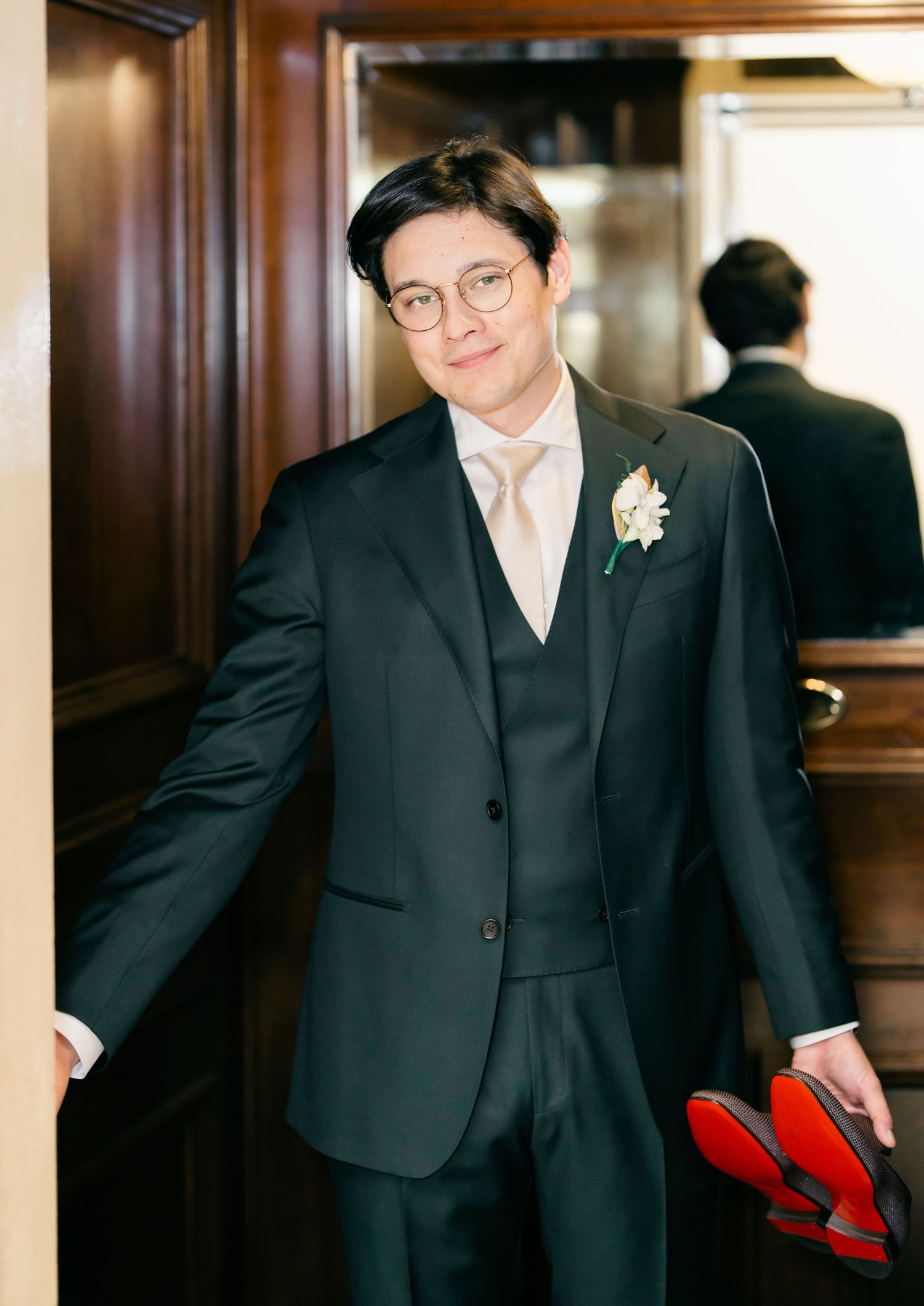 A groom in a classic black three-piece suit with a light tie and boutonniere poses elegantly near a mirrored wall.