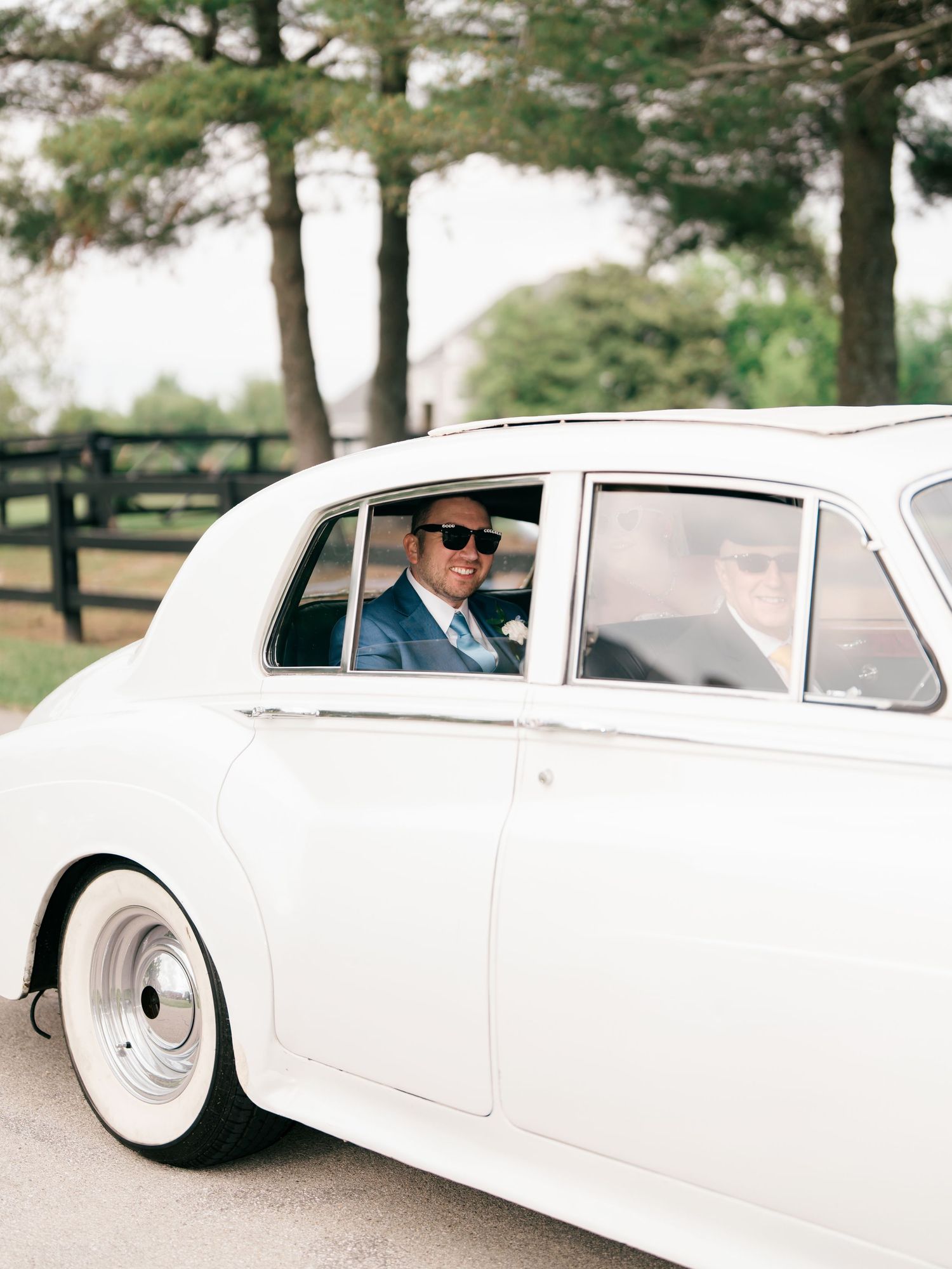 Groom in blue suit and sunglasses arriving in a classic white Rolls-Royce on a tree-lined driveway.