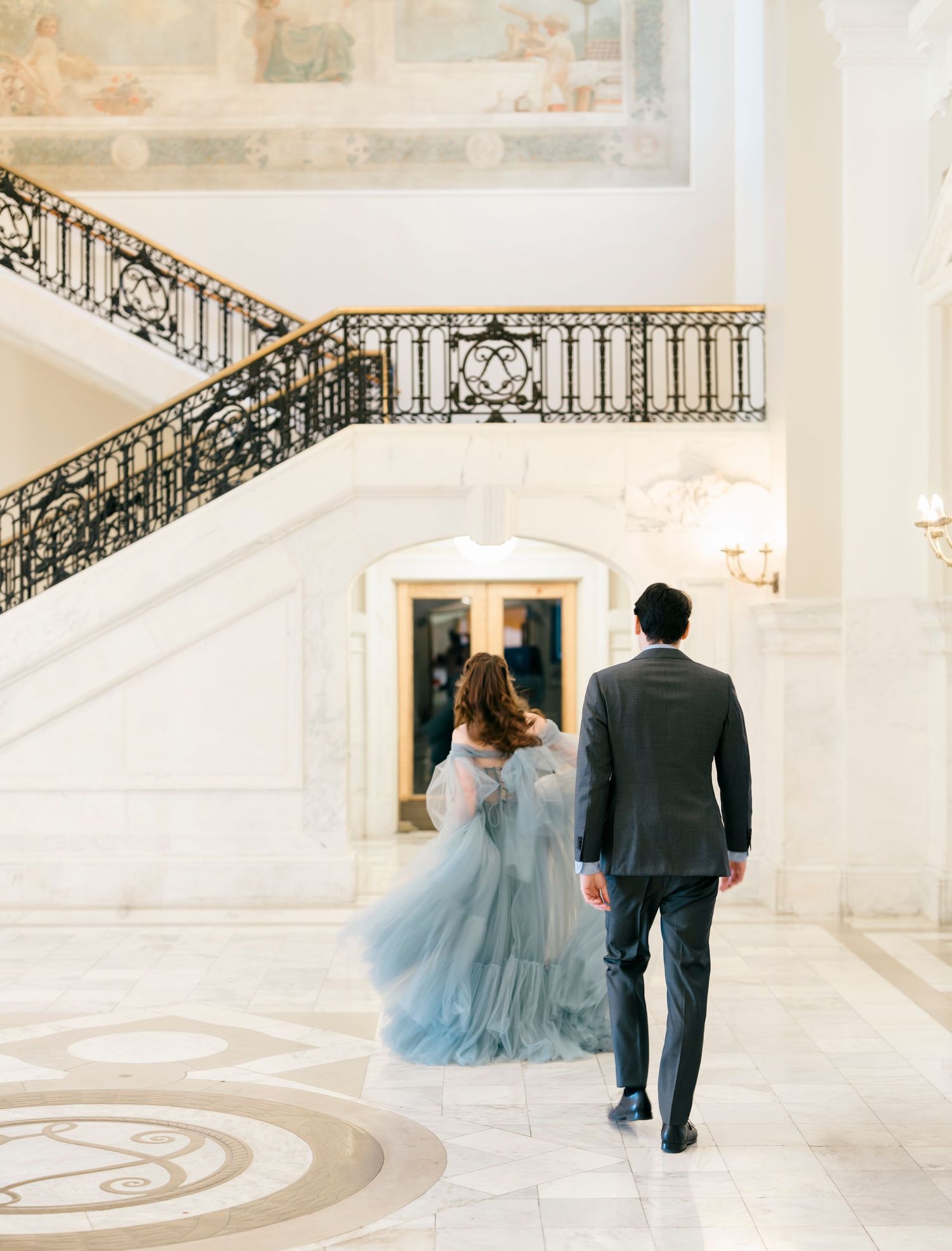 A couple in formal attire walks up an elegant white marble staircase with ornate black railings in a grand venue.