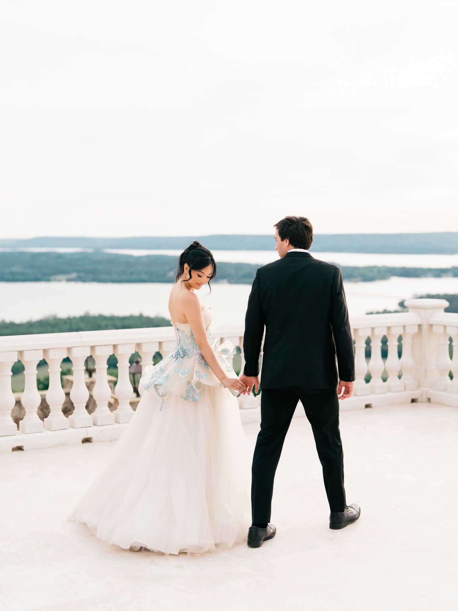 Bride and groom hold hands on white balcony terrace overlooking scenic lake and green hills.