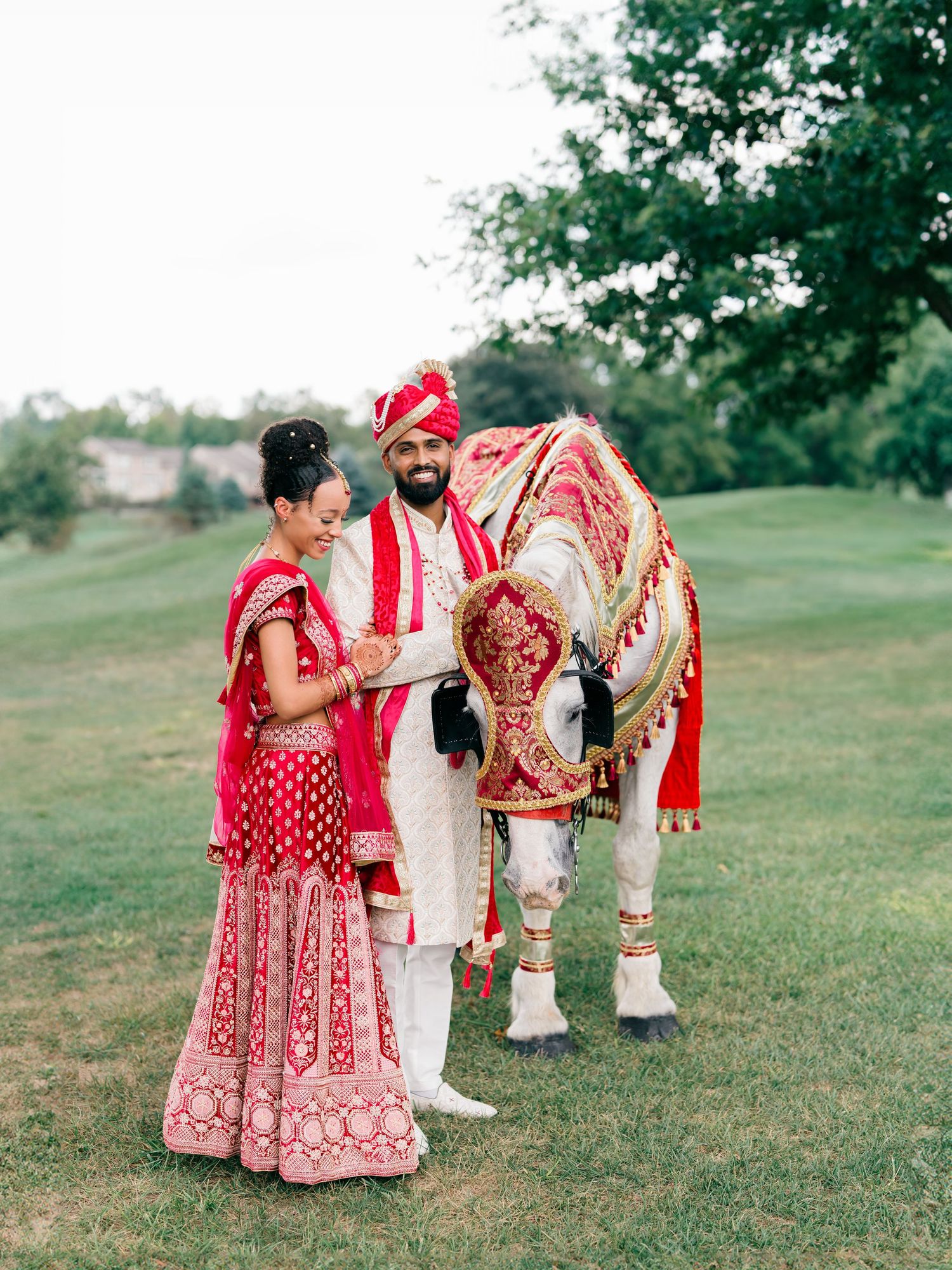 Indian bride and groom in red traditional attire stand beside a decorated white horse on a lush green lawn.
