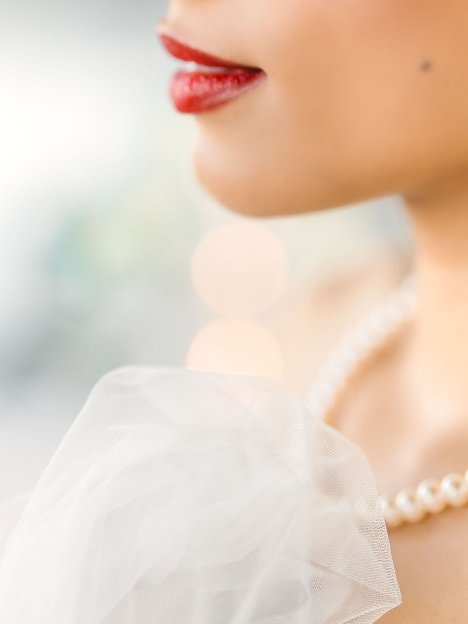 Close up detail of a Bride with delicate white tulle veil fabric, red lipstick, and pearl necklace, creating an ethereal bridal aesthetic.