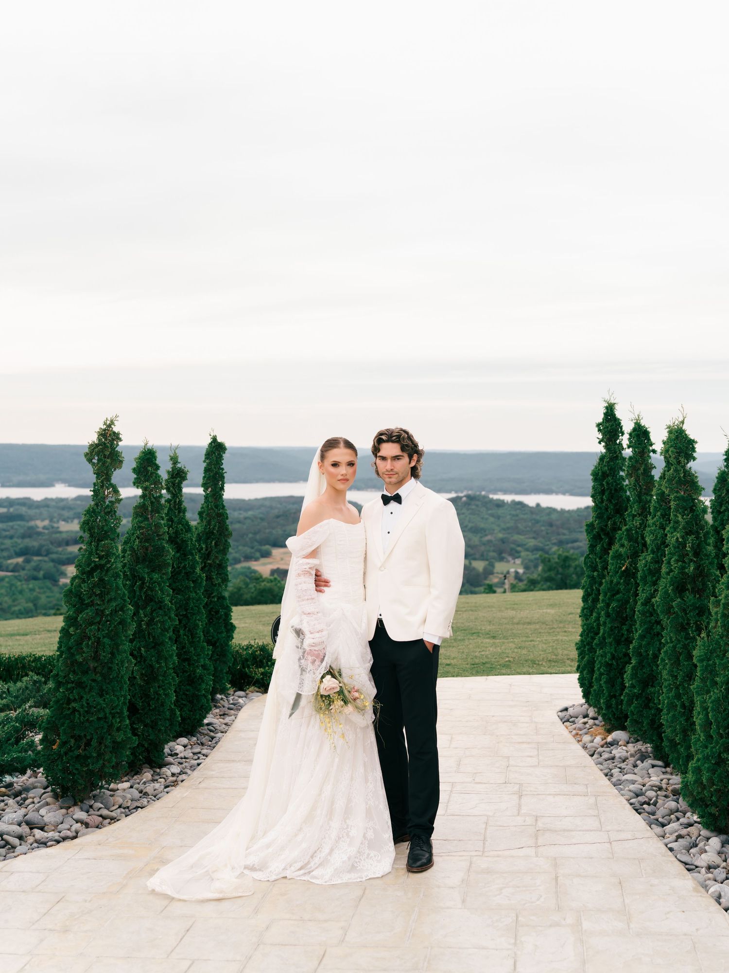 Bride and groom pose on stone path flanked by tall cypress trees with panoramic lake views.