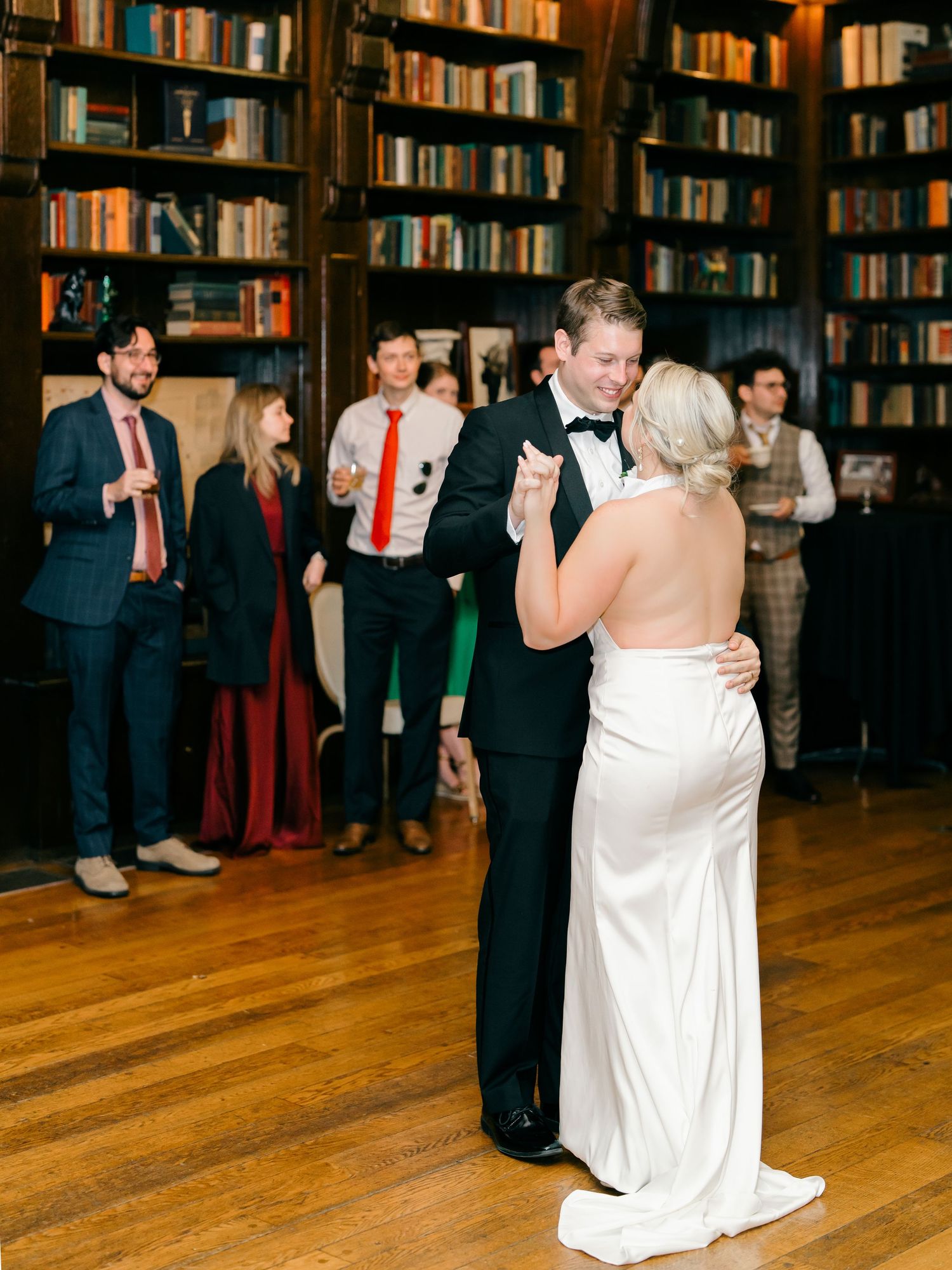 A couple shares their first dance in a library wedding venue with dark wood bookshelves and guests watching in the background.