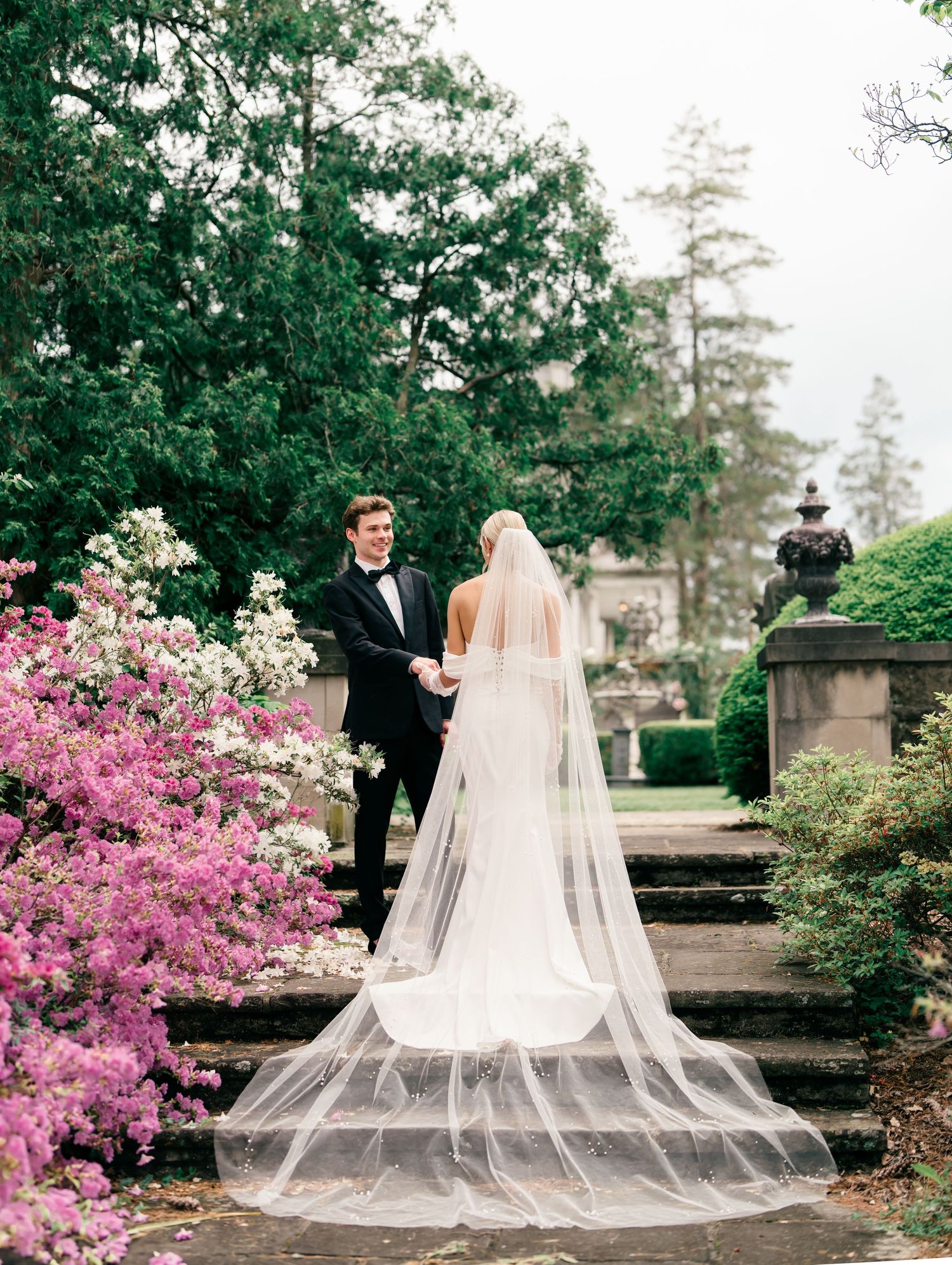 Garden wedding portrait on stone steps with dramatic cathedral veil and pink blooming azaleas.