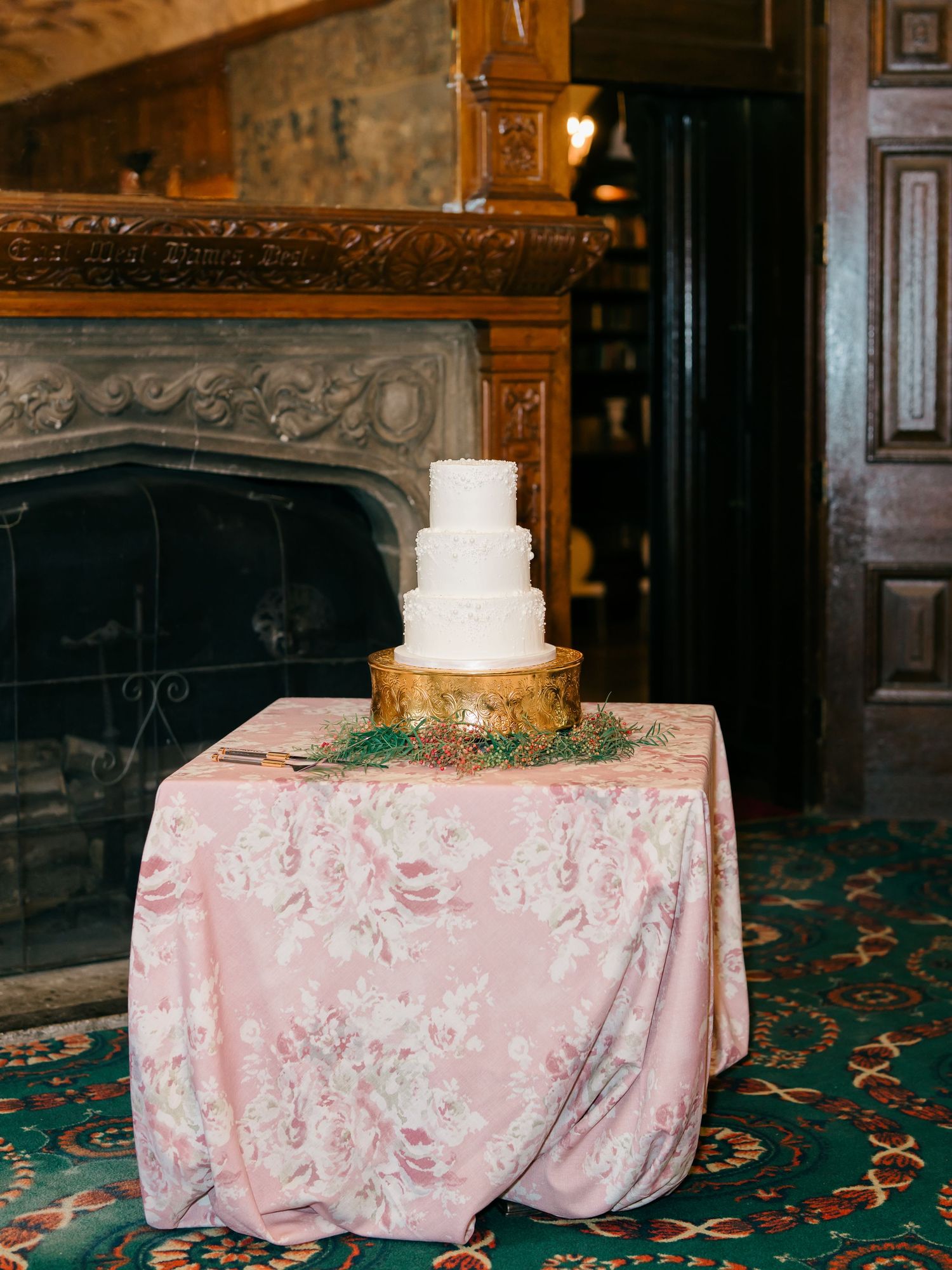 A wedding cake displayed on a pink floral tablecloth in an elegant wood-paneled room with ornate details.