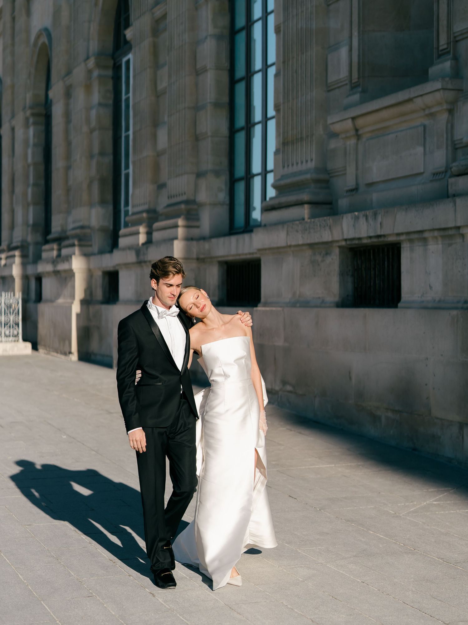 A couple in wedding attire walks along a stone building exterior casting dramatic shadows at sunrise in Paris, France.