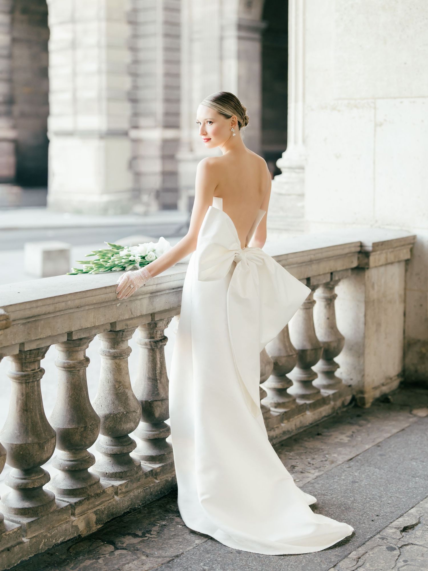 Elegant bride in white gown poses gracefully along historic stone balustrade in romantic outdoor setting at The Louvre in Paris, France.