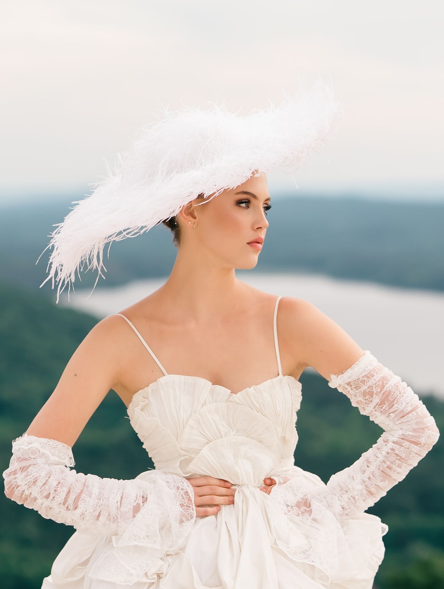 Bride in white feather hat, lace gloves, and strapless gown poses outdoors with scenic green hills and lake behind her.